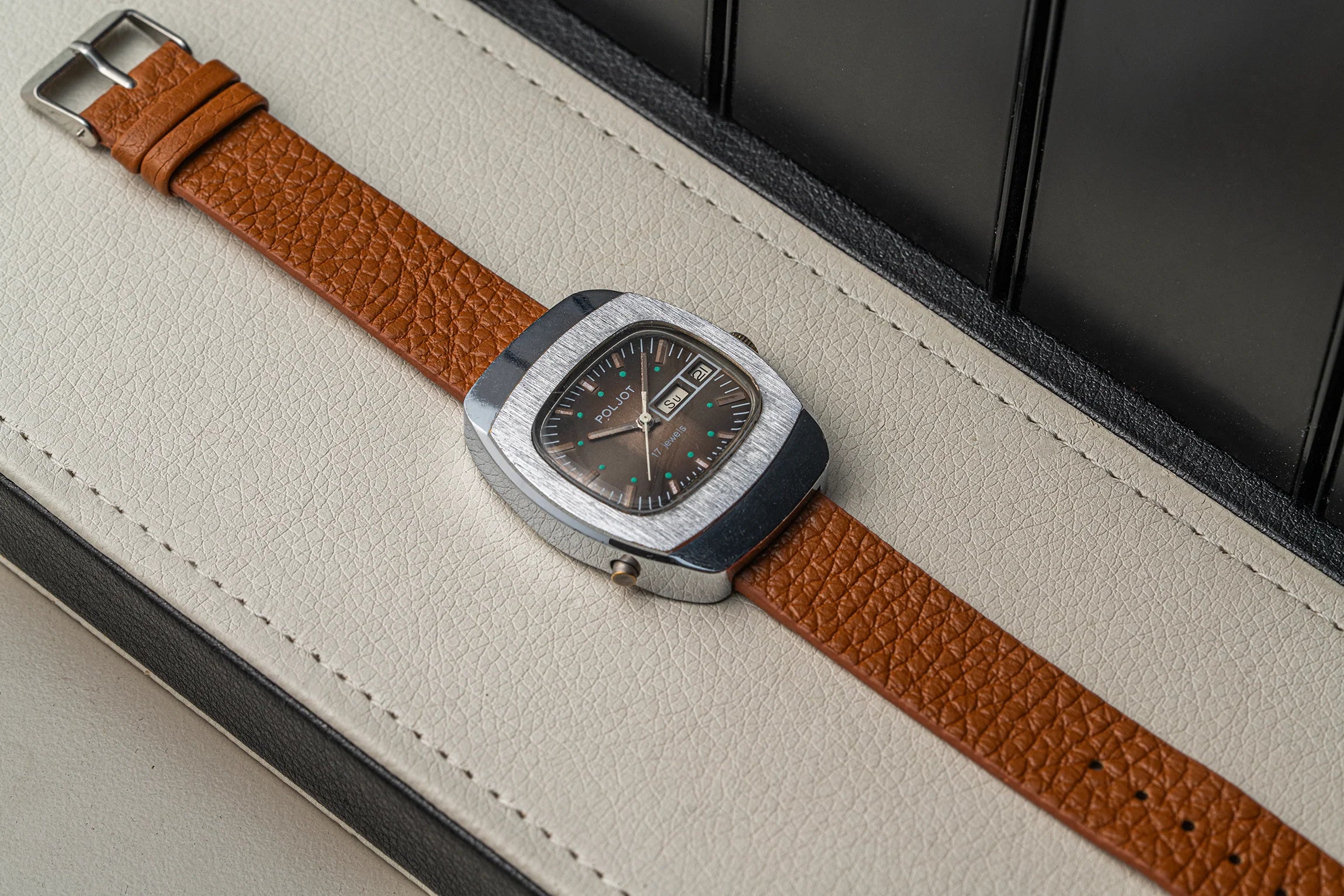 A vintage-style wristwatch Poljot with a silver-toned square case, dark dial, and a textured brown leather strap, resting diagonally on a cream-colored leather surface.