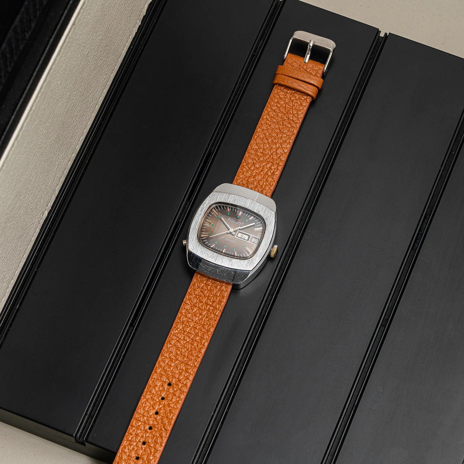 A vintage-style wristwatch Poljot with a textured orange-brown leather strap and a silver-toned cushion-shaped case, displayed diagonally on a black presentation box.