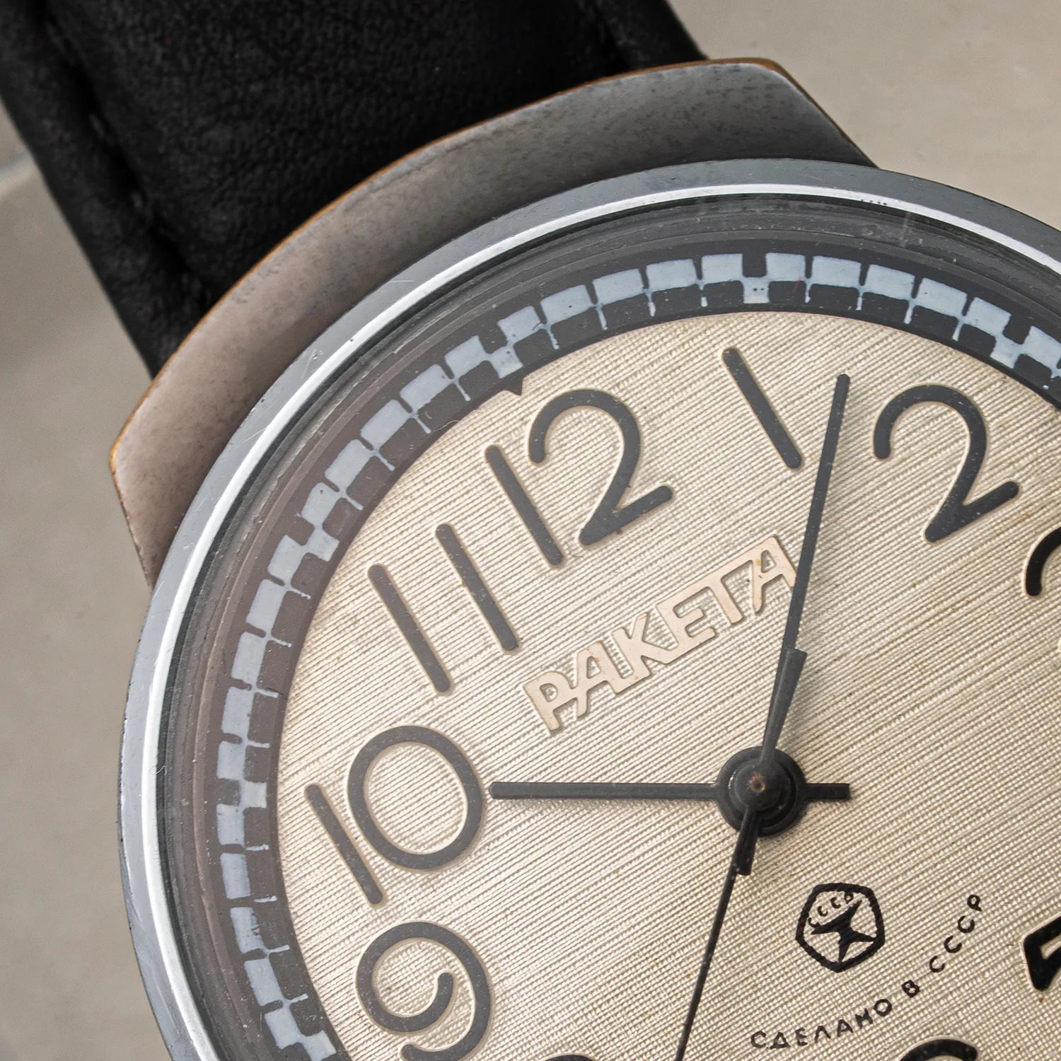 Close-up of a vintage-style wristwatch Raketa Big Numerals with a textured beige dial, bold black Arabic numerals, black hands, a silver-toned bezel, and a black leather strap.