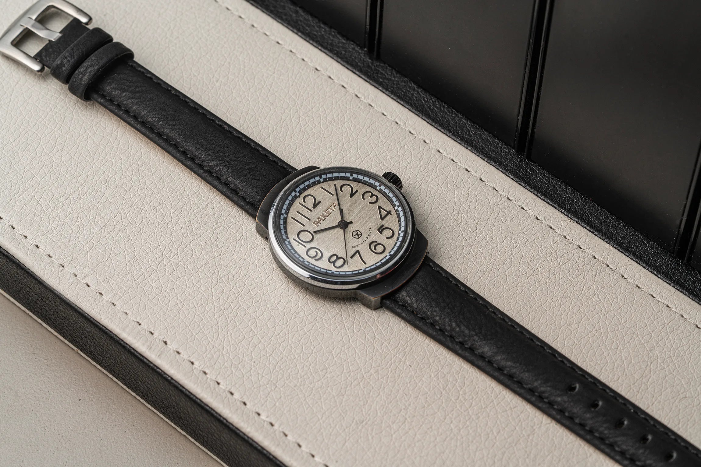 A close-up of a wristwatch Raketa Big Numerals with a black leather strap and a silver-toned round case, resting on a cream-colored textured surface with a dark background.
