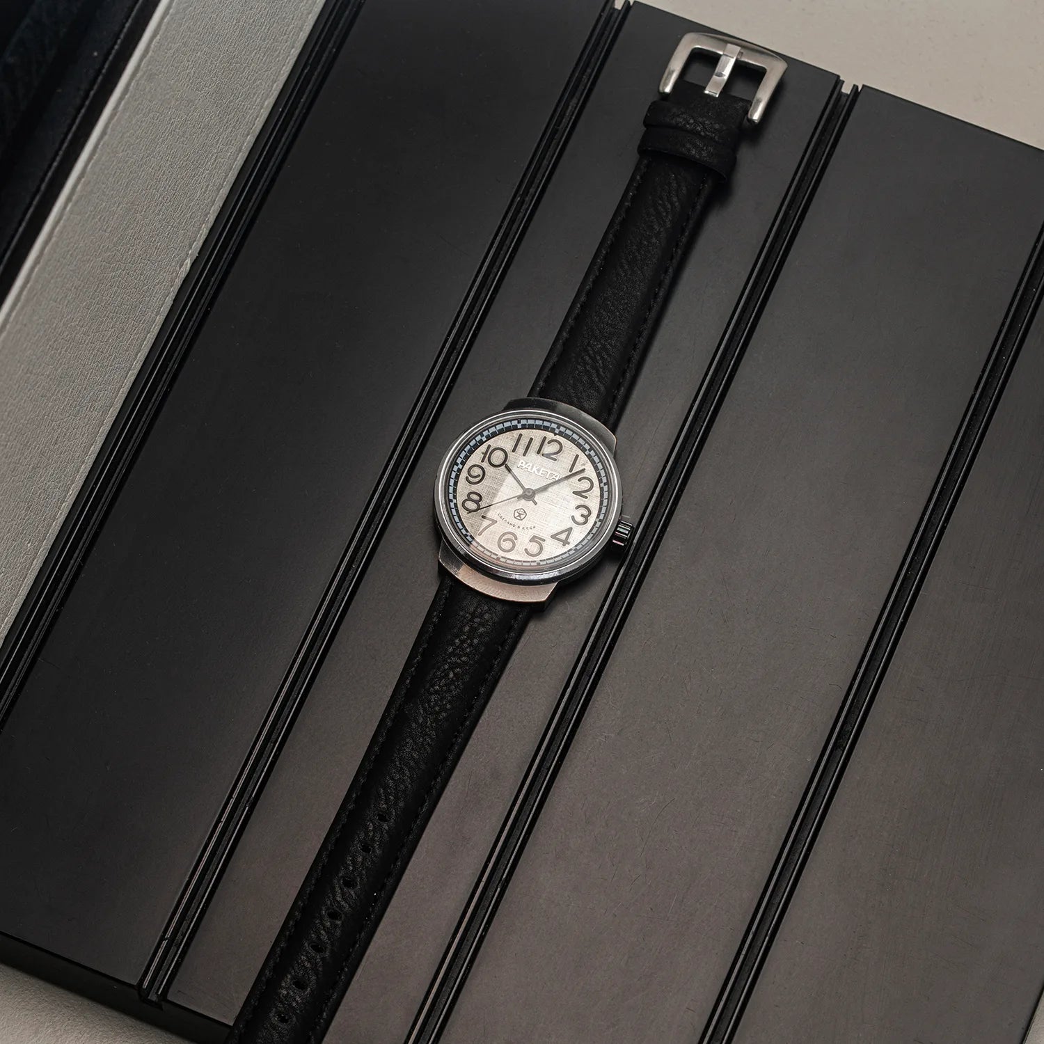 A wristwatch Raketa Big Numerals with a black textured leather strap and a silver-toned round dial, displayed diagonally on a dark gray textured surface.