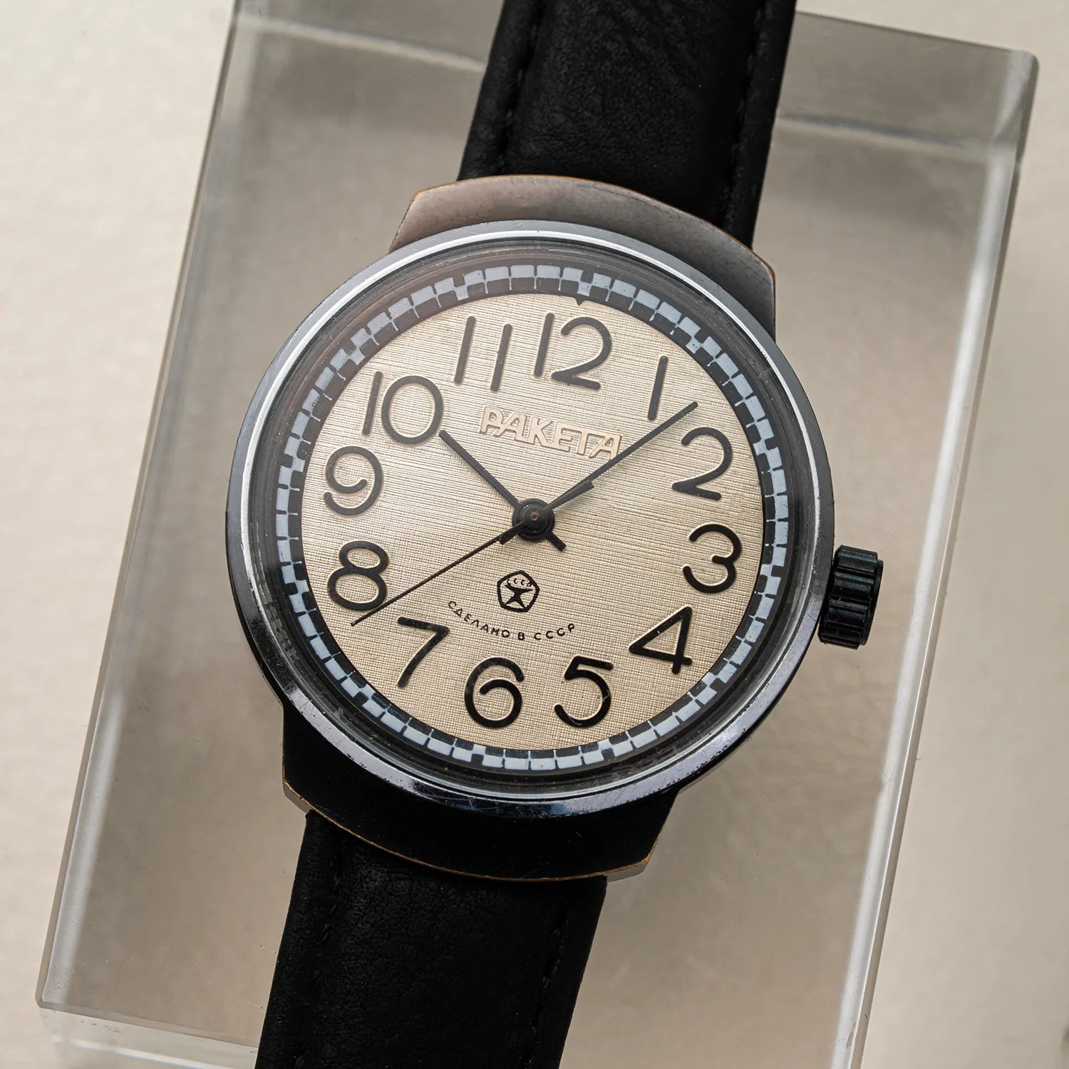 A vintage-style Raketa Big Numerals watch with a black leather strap, a brushed silver-tone case, and a cream-colored dial with black Arabic numerals, displayed on a clear acrylic stand.