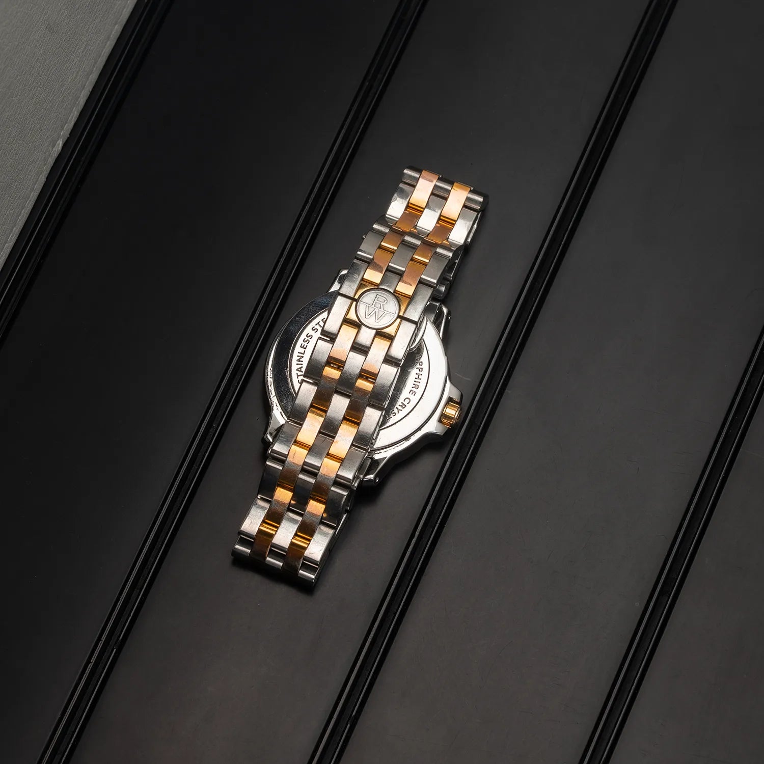 A two-tone stainless steel watch Raymond Weil Tango 5599 with a silver and gold link bracelet, shown from the back on a dark textured background.