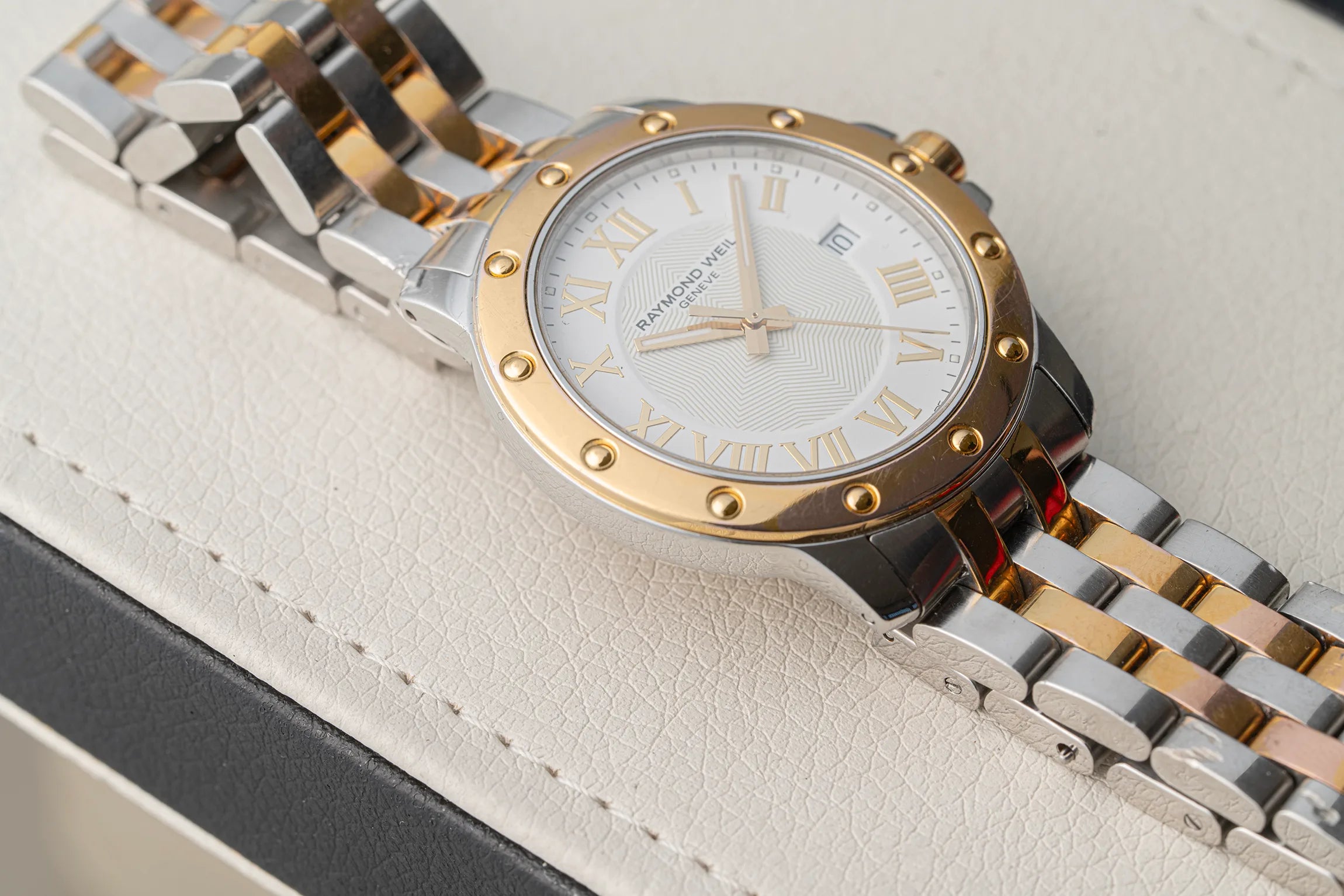 A close-up of a two-tone stainless steel and gold watch Raymond Weil Tango 5599 with a white dial, Roman numeral hour markers, and a screw-head bezel, resting on a textured cream-colored leather surface.