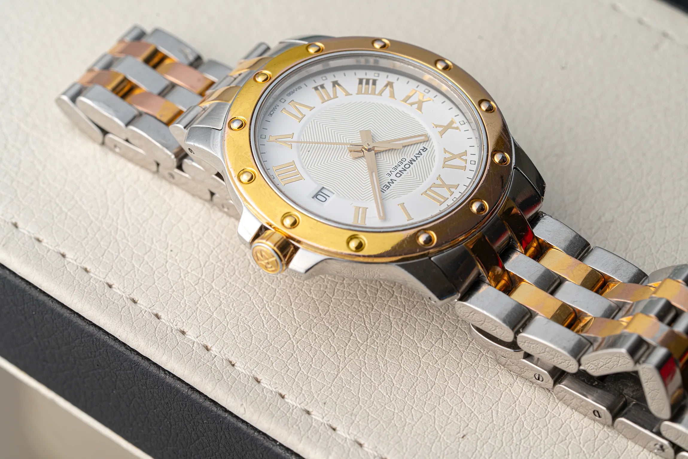A close-up of a Raymond Weil Tango 5599 watch with a two-tone silver and gold stainless steel link bracelet and a gold bezel, resting on a textured cream-colored leather surface.