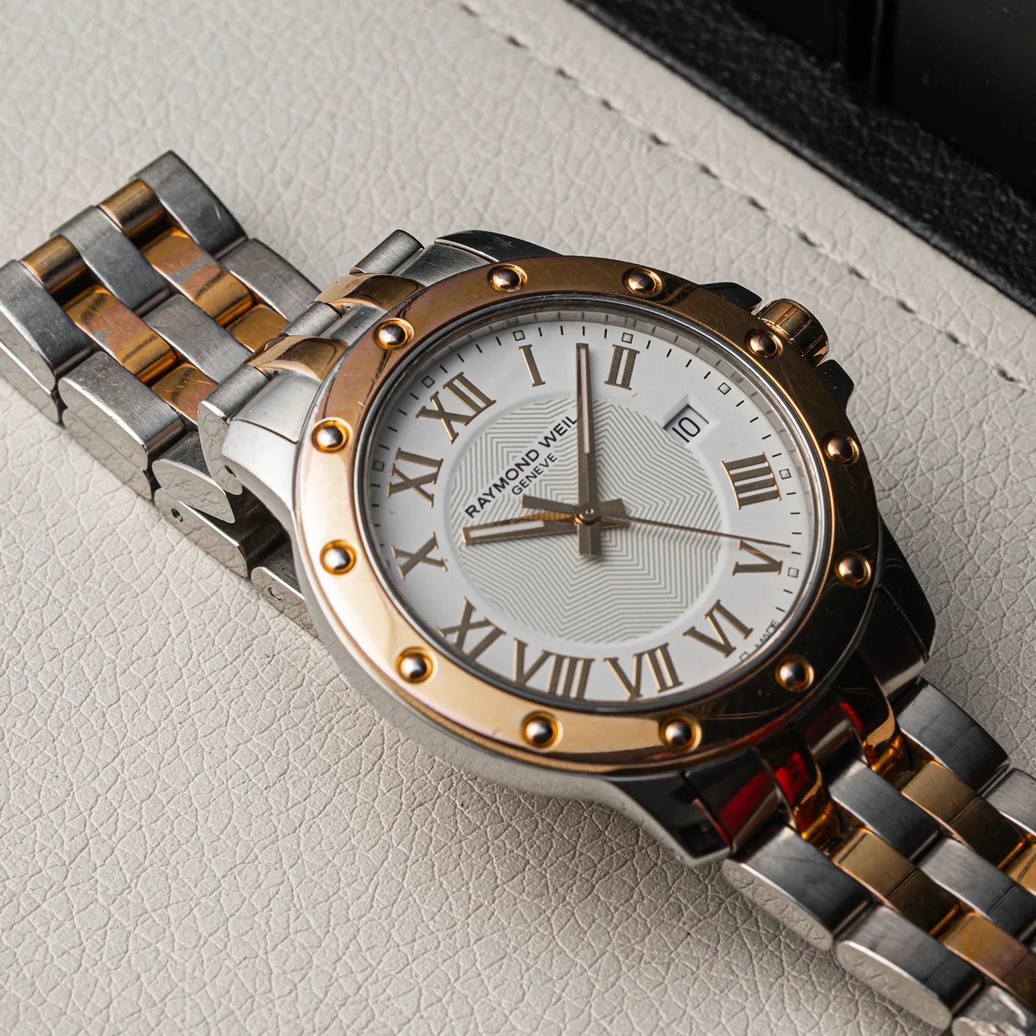 A close-up of a Raymond Weil Tango 5599 wristwatch with a two-tone silver and gold-tone metal bracelet, a gold-tone bezel, and a white dial with Roman numerals, resting on a textured light beige leather surface.