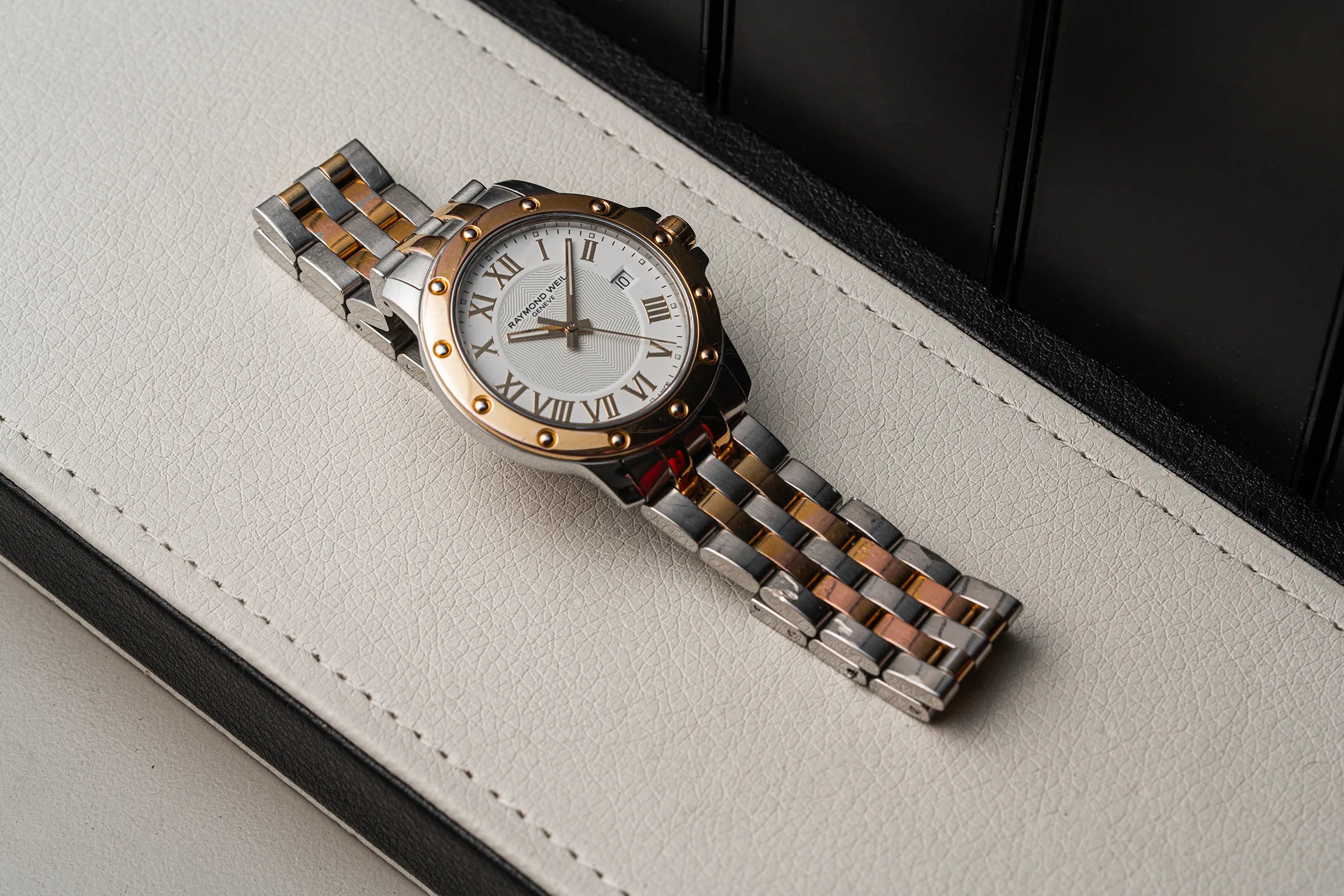 A two-tone stainless steel and rose gold watch Raymond Weil Tango 5599 with a white dial and Roman numerals, resting diagonally on a textured off-white leather surface.