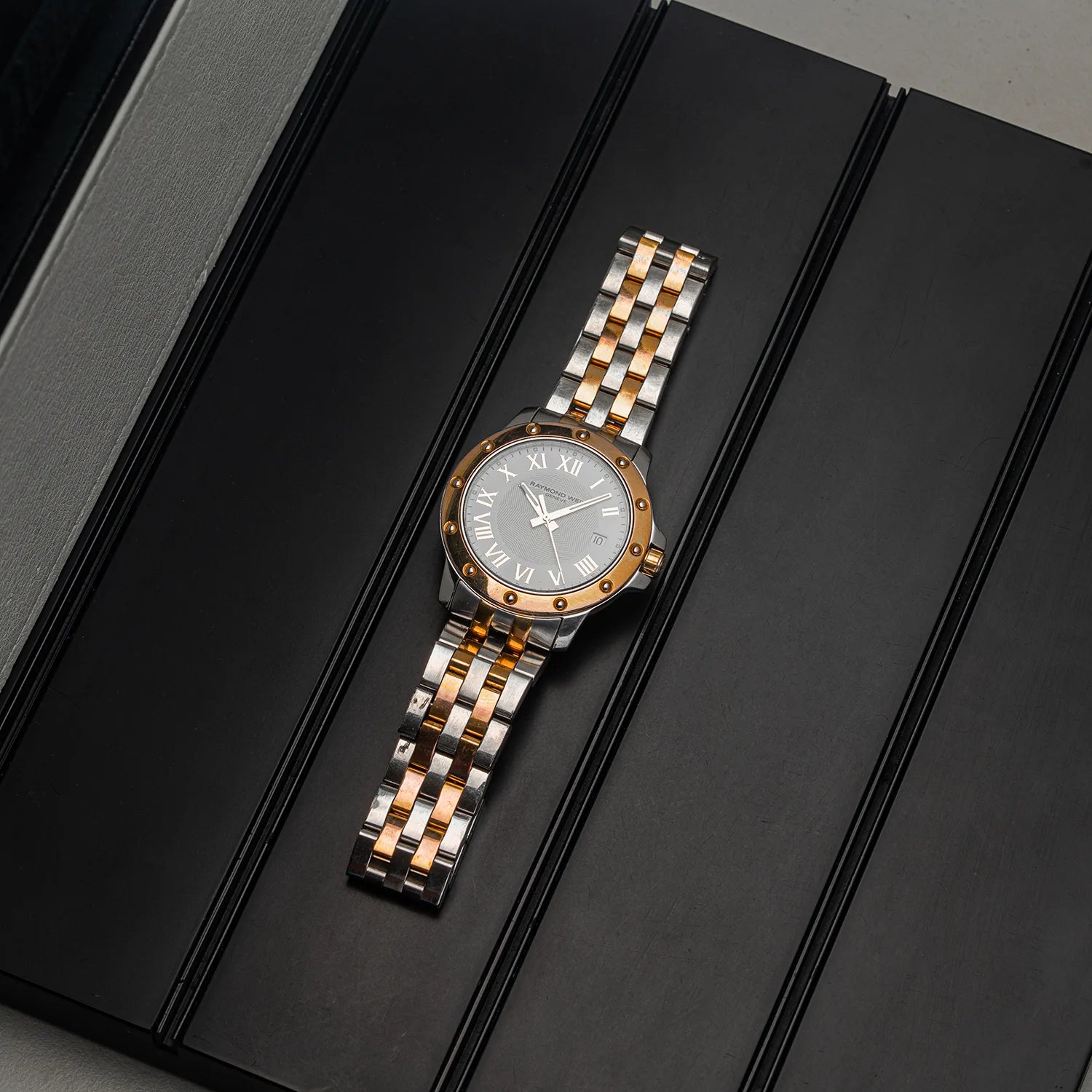 A two-tone stainless steel and rose gold wristwatch Raymond Weil Tango 5599 with a silver dial and Roman numeral hour markers, resting diagonally on a black textured surface.