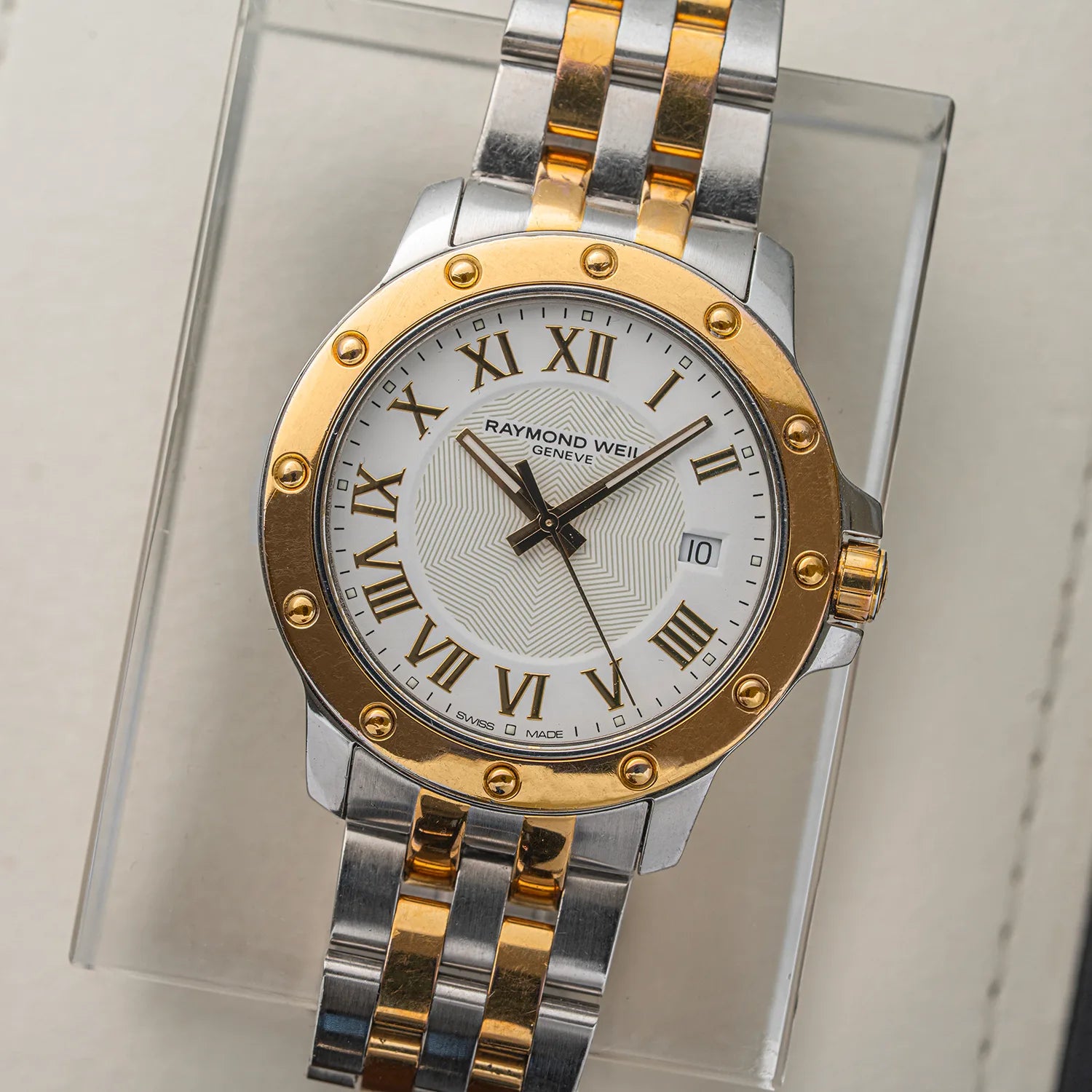 A Raymond Weil Tango 5599 watch with a two-tone silver and gold stainless steel bracelet, a gold bezel, and a white dial with gold Roman numerals, displayed on a clear acrylic stand.