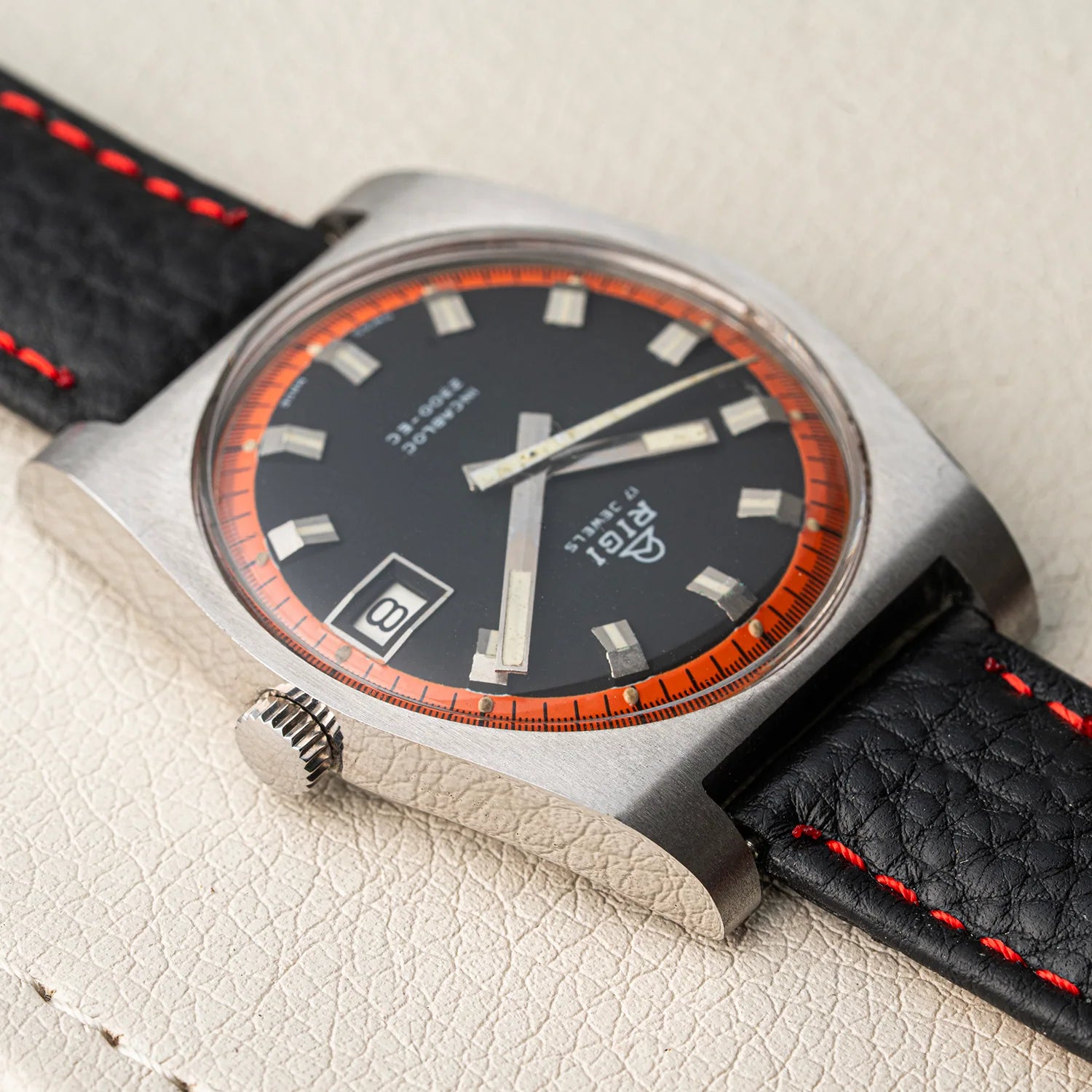A close-up of a vintage-style wristwatch Rigi 2300-EC with a brushed silver-tone case, black dial, orange bezel accents, and a black leather strap with red stitching, resting on a light textured surface.