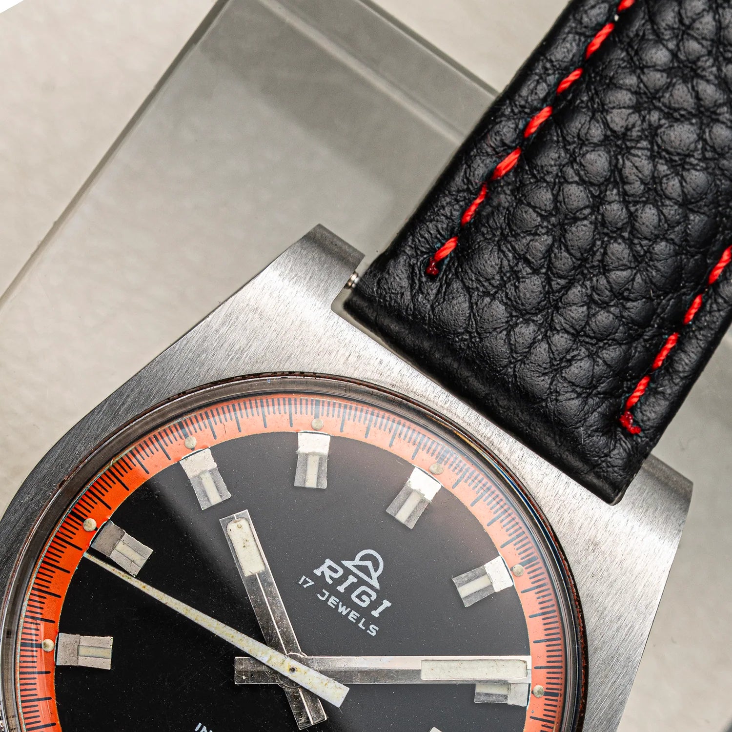 Close-up of a wristwatch Rigi 2300-EC with a brushed silver-tone case, black dial with orange ring, silver hour markers and hands, and a black textured leather strap with red stitching.
