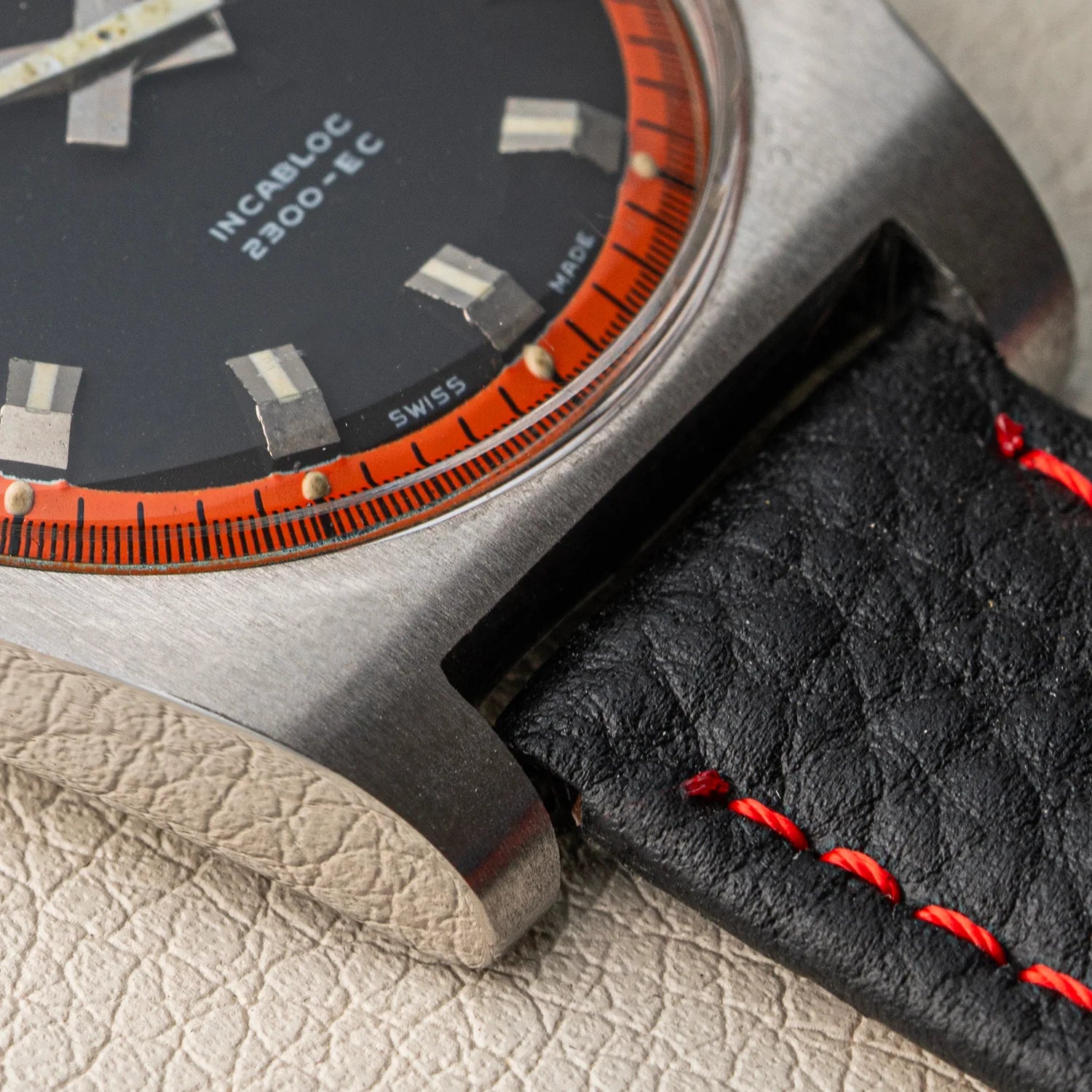 Close-up of a wristwatch Rigi 2300-EC with a brushed metal case, black dial, orange outer ring, and a black leather strap with red stitching, resting on a light textured surface.