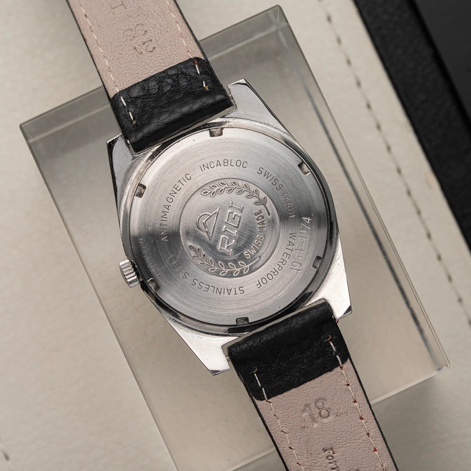 Close-up of the back of a wristwatch Rigi 2300-EC with a stainless steel case and a light beige leather strap, resting on a clear display box. The watch case is engraved with 'A.