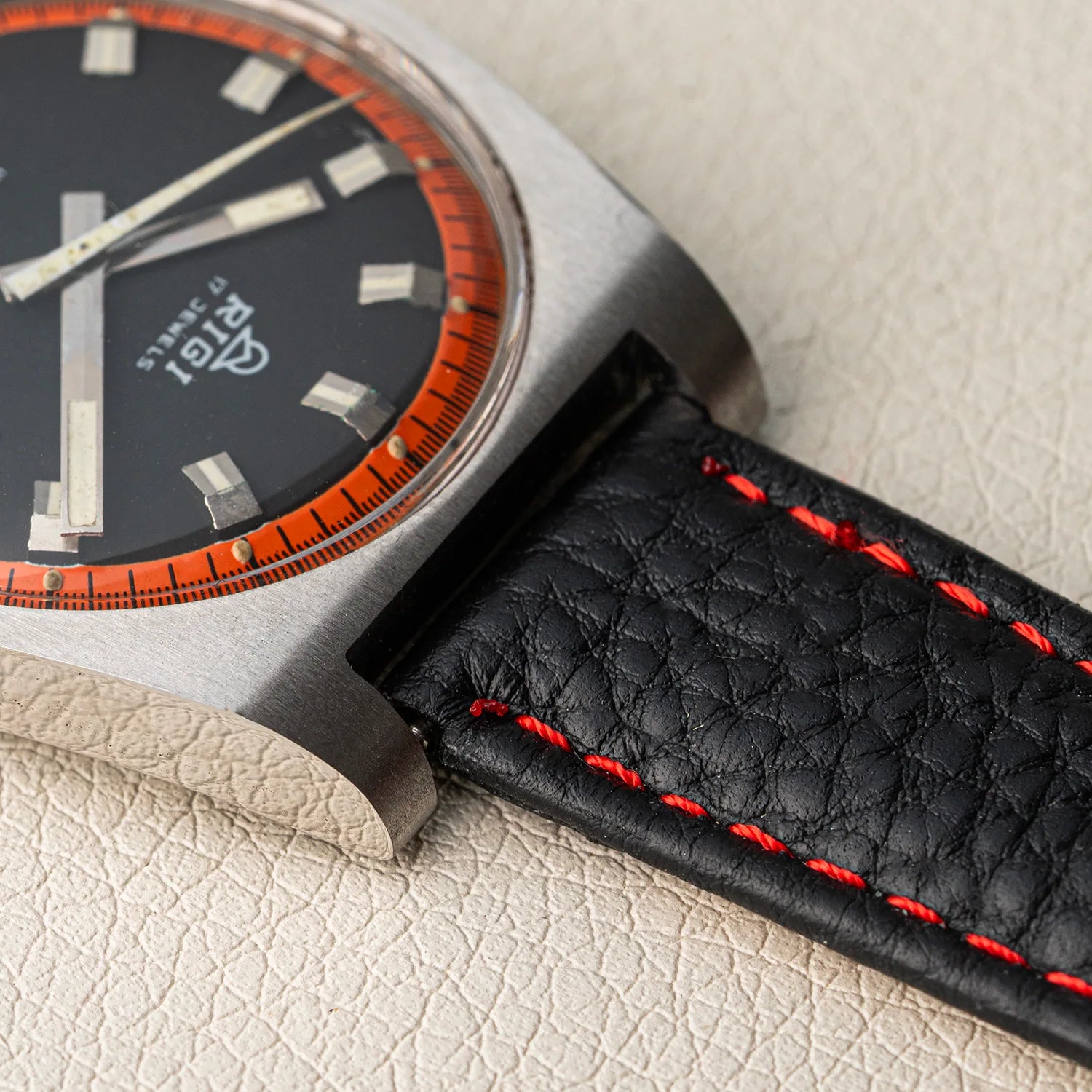 Close-up of a wristwatch Rigi 2300-EC with a black dial, orange bezel markings, and a black leather strap with red stitching, resting on a textured beige surface.