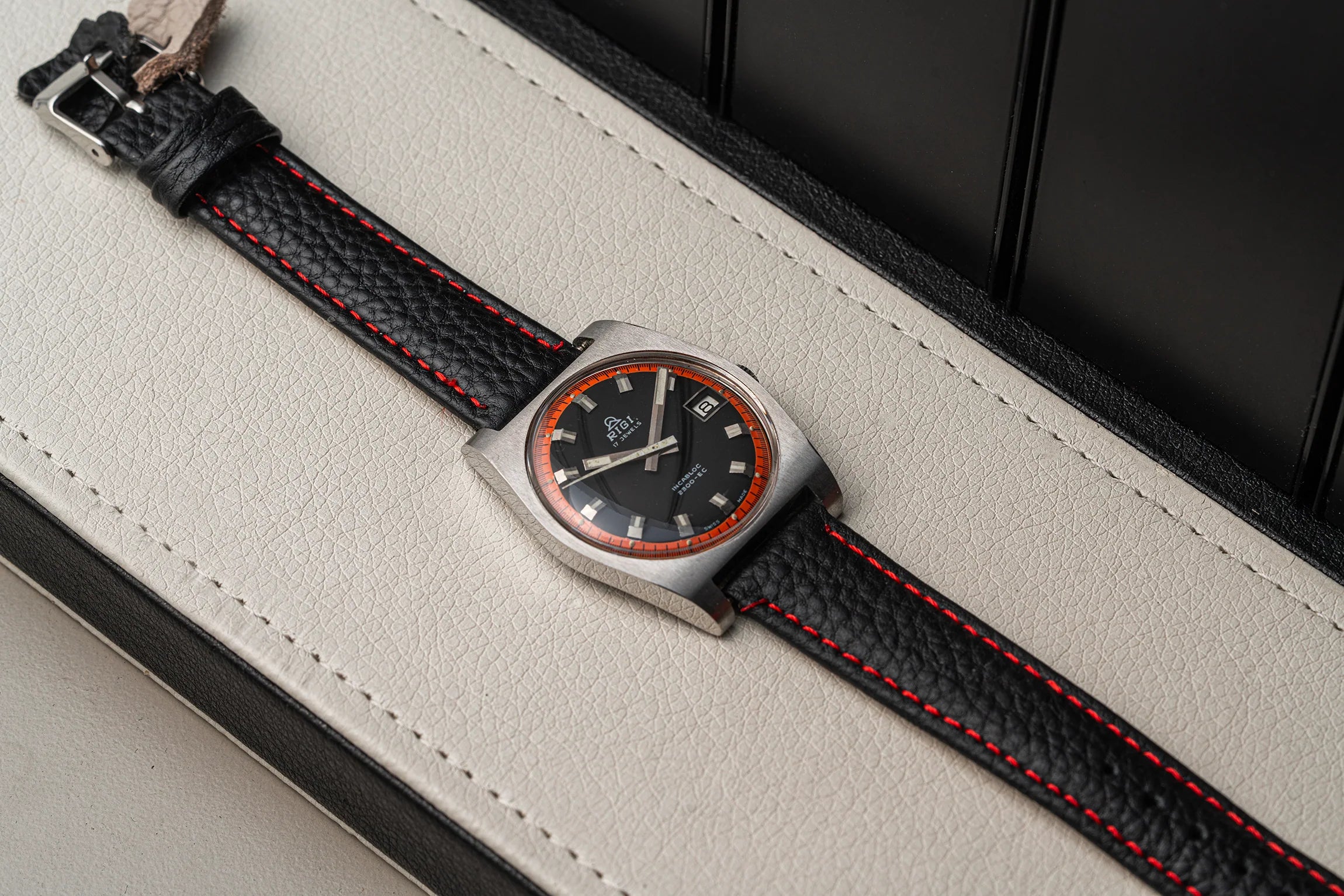 A wristwatch Rigi 2300-EC with a silver-toned case, black dial, and black leather strap with red stitching, resting diagonally on a cream-colored textured surface.