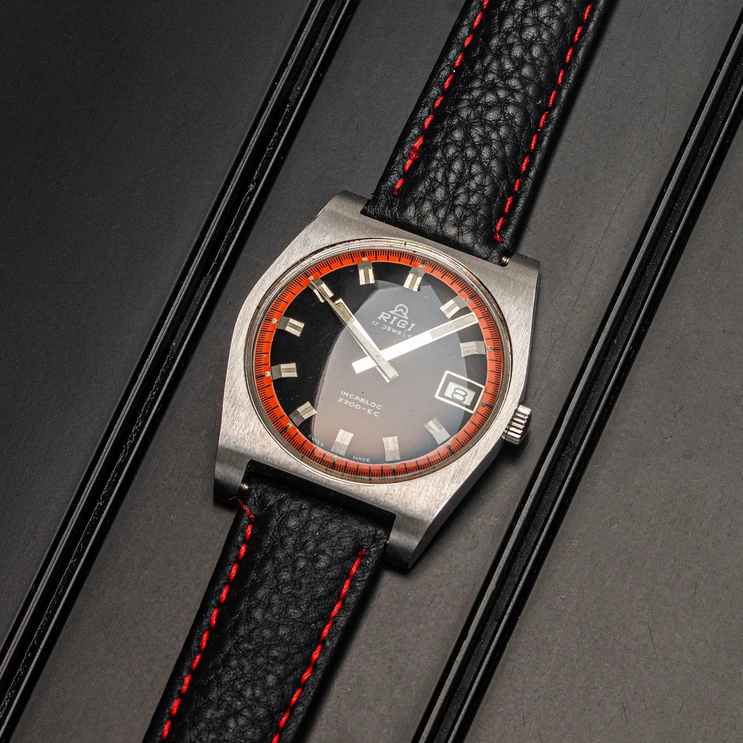 A vintage-style wristwatch Rigi 2300-EC with a brushed silver-tone case, black dial, orange ring around the bezel, and a black textured leather strap with red stitching, displayed diagonally on a dark textured background.