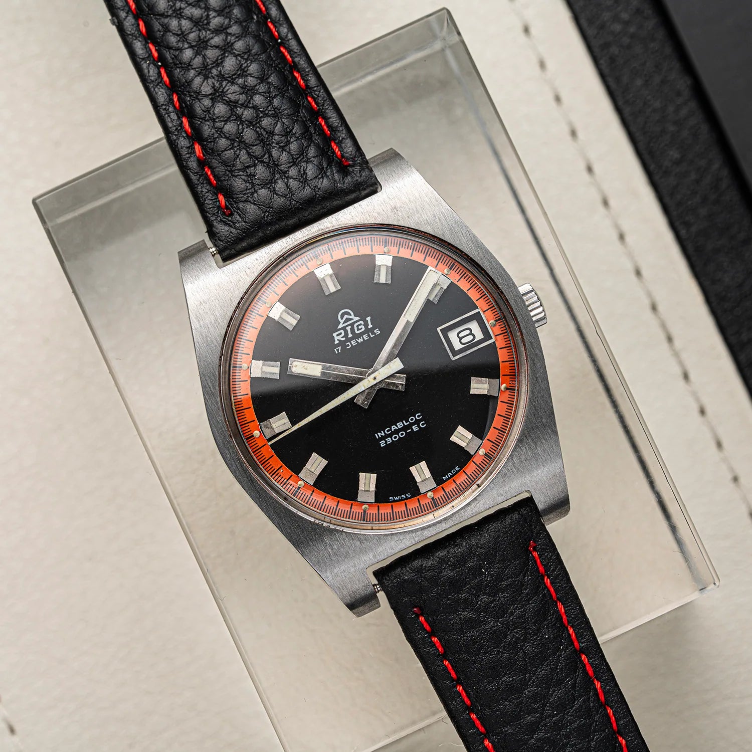 A vintage-style wristwatch Rigi 2300-EC with a brushed silver-tone case, black dial, orange ring around the bezel, and a black textured leather strap with red stitching, displayed on a clear box.