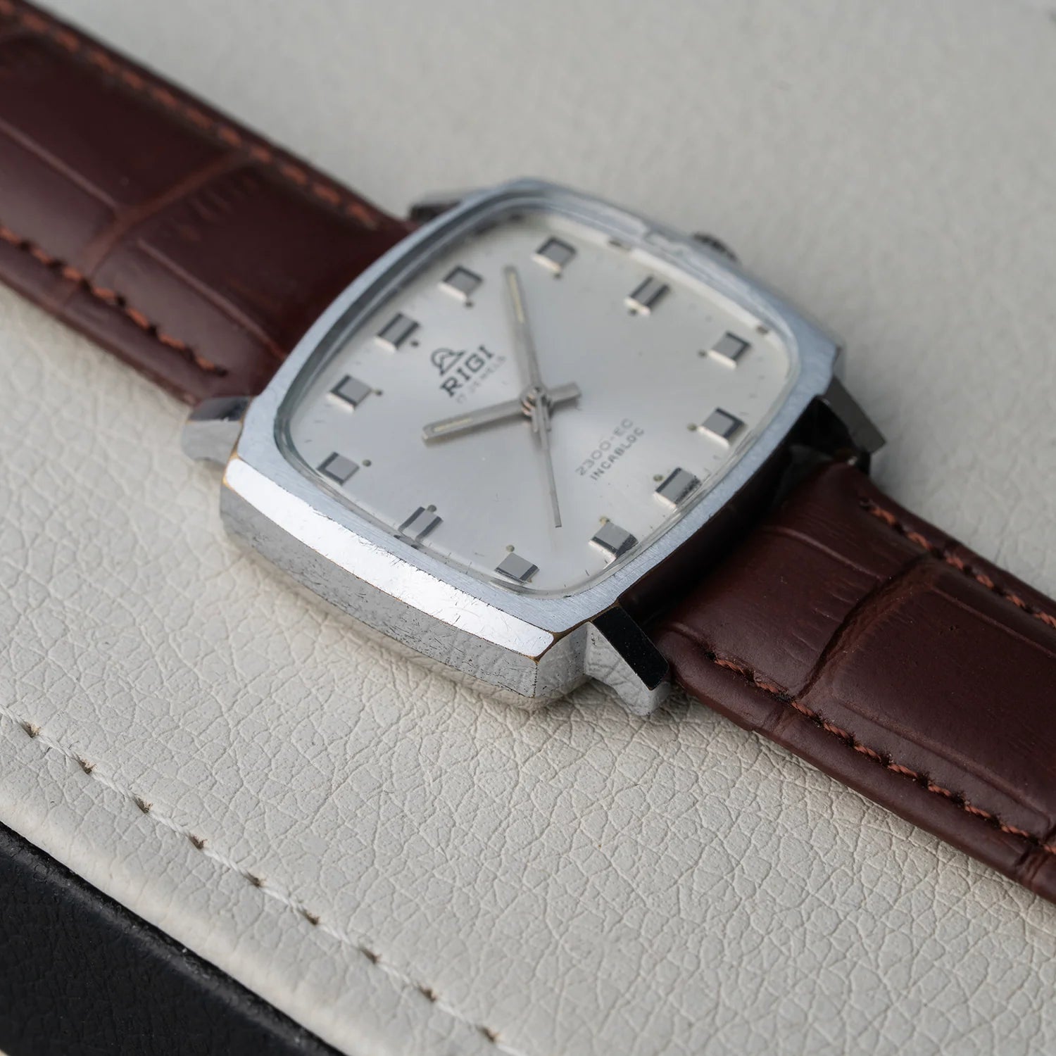 A vintage-style wristwatch Rigi 2300-EC, Cal. AS 1950 with a silver-toned square case, silver dial, and brown leather strap, photographed close-up on a light textured surface.