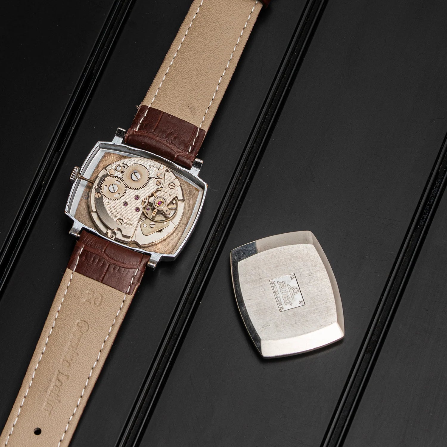 A luxury watch with a rectangular silver-toned case, featuring a visible mechanical movement AS 1950 through a polished backplate and a beige leather strap with brown leather accents, shown alongside its detached silver backplate on a black textured background.