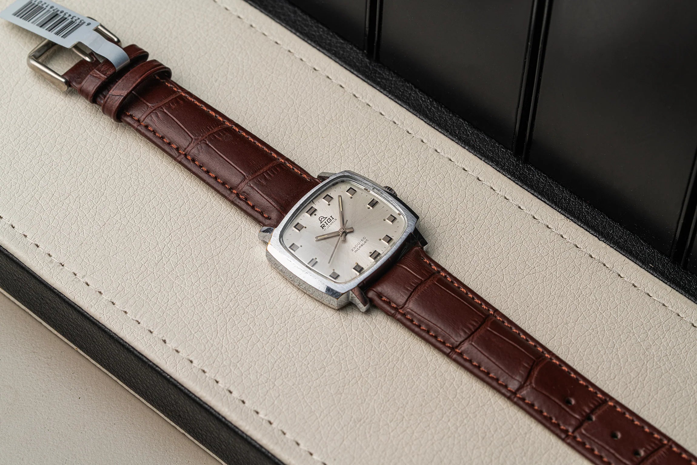 A vintage-style wristwatch Rigi 2300-EC, Cal. AS 1950 with a brown alligator-embossed leather strap and a silver-toned rectangular case, resting diagonally on a cream-colored textured leather surface.