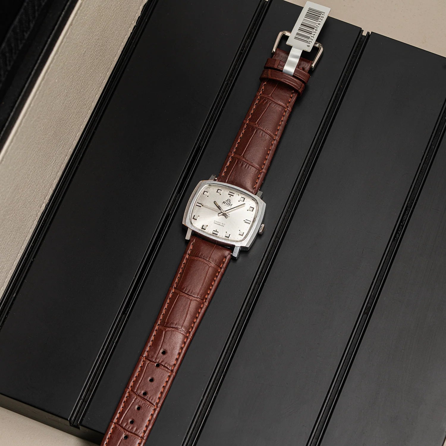 A vintage-style wristwatch Rigi 2300-EC, Cal. AS 1950 with a brown alligator leather strap and a silver-toned rectangular case, displayed diagonally on a black presentation box.