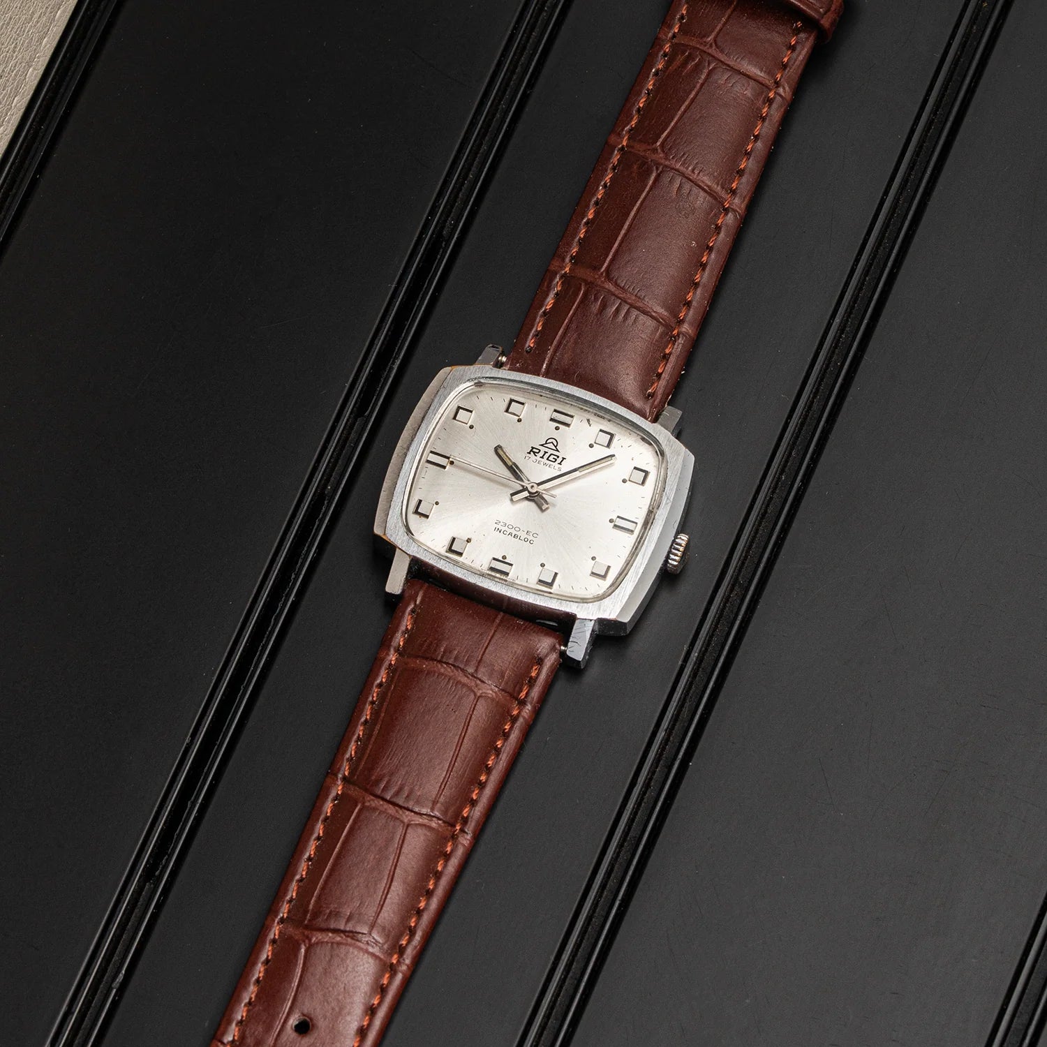 A vintage-style wristwatch with a silver-toned rectangular case, white dial, and brown alligator-embossed leather strap, photographed on a dark textured surface.