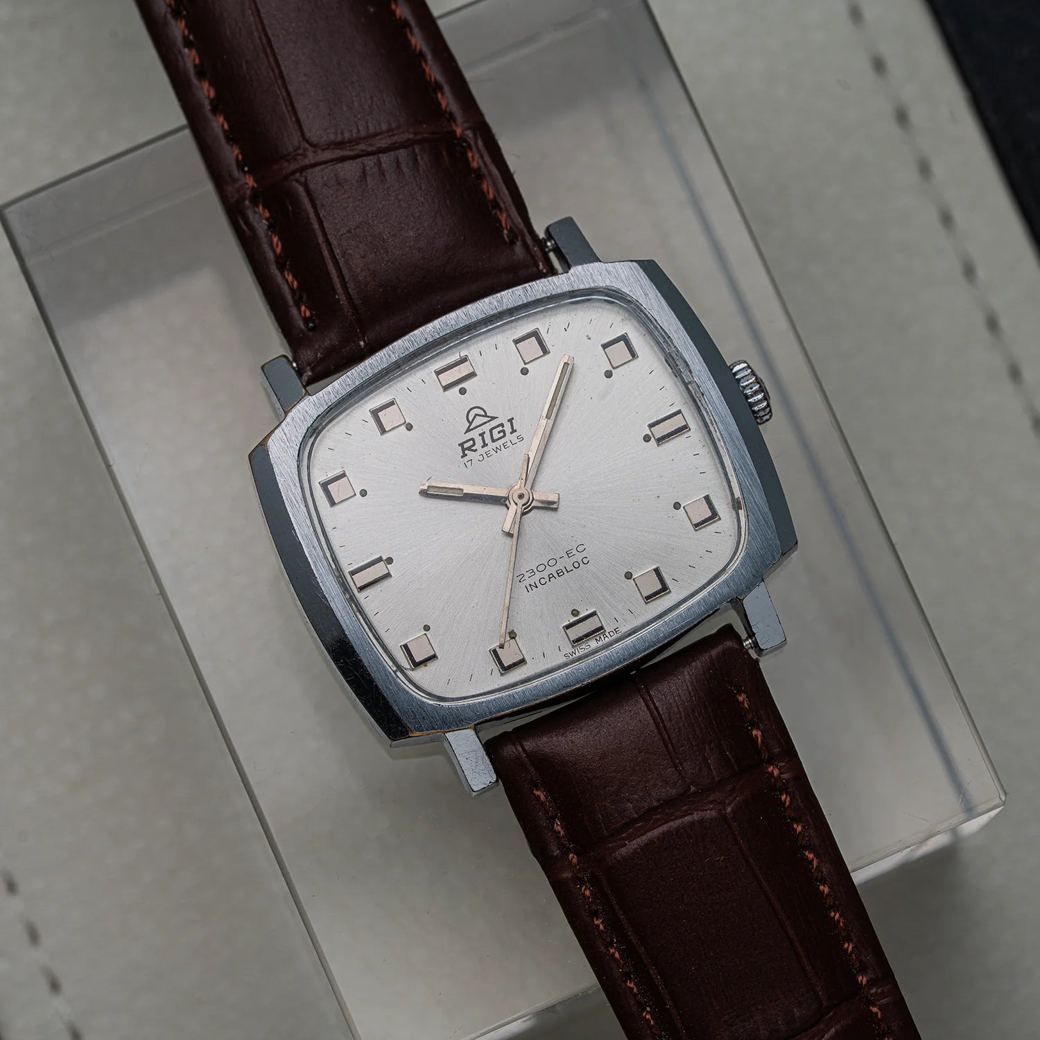 A vintage RIG! wristwatch with a silver-toned square case, white dial, and dark brown leather strap, resting on its light gray presentation box.