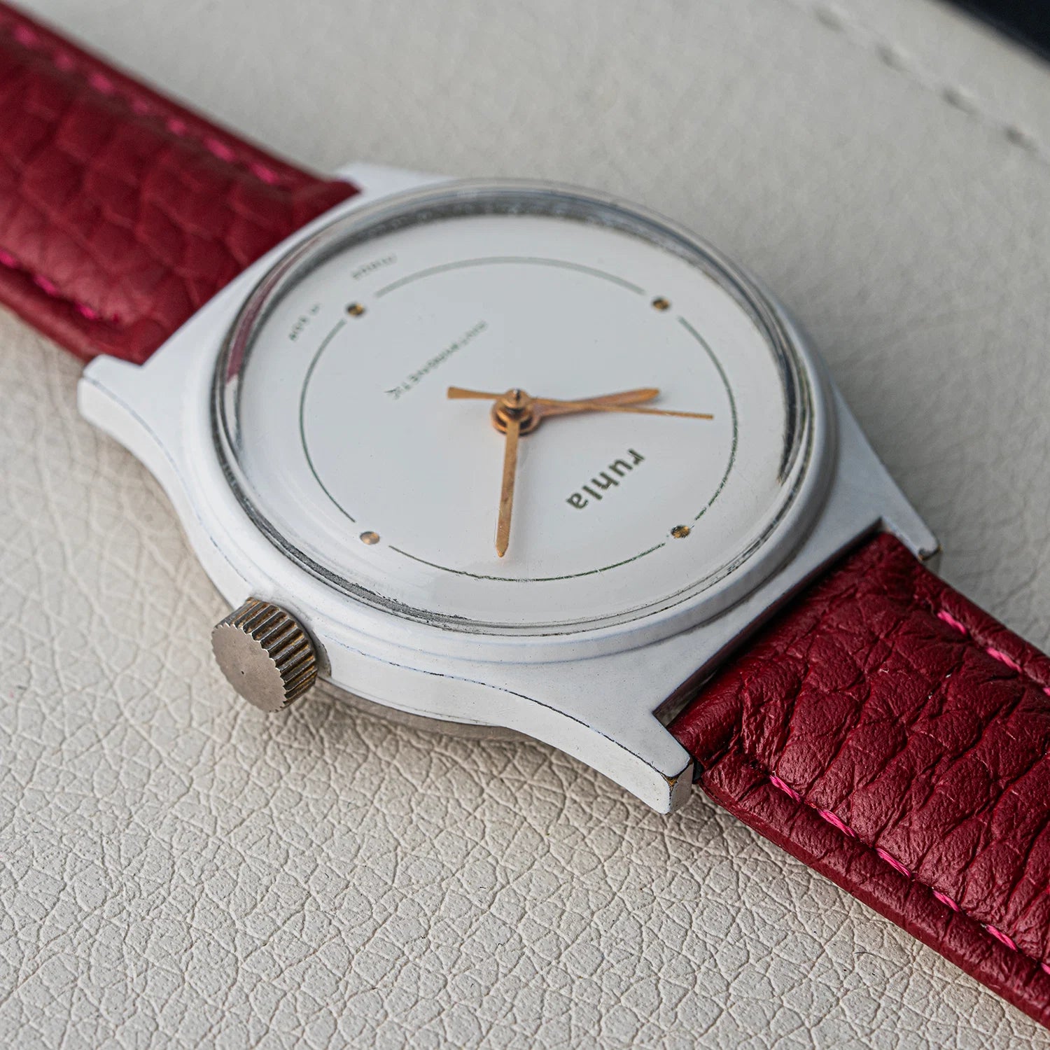 A close-up of a vintage-style wristwatch    Ruhla, Cal. UMF 24-42with a white dial, gold-tone hands and markers, a silver-tone case, and a textured deep red leather strap on a light textured surface.