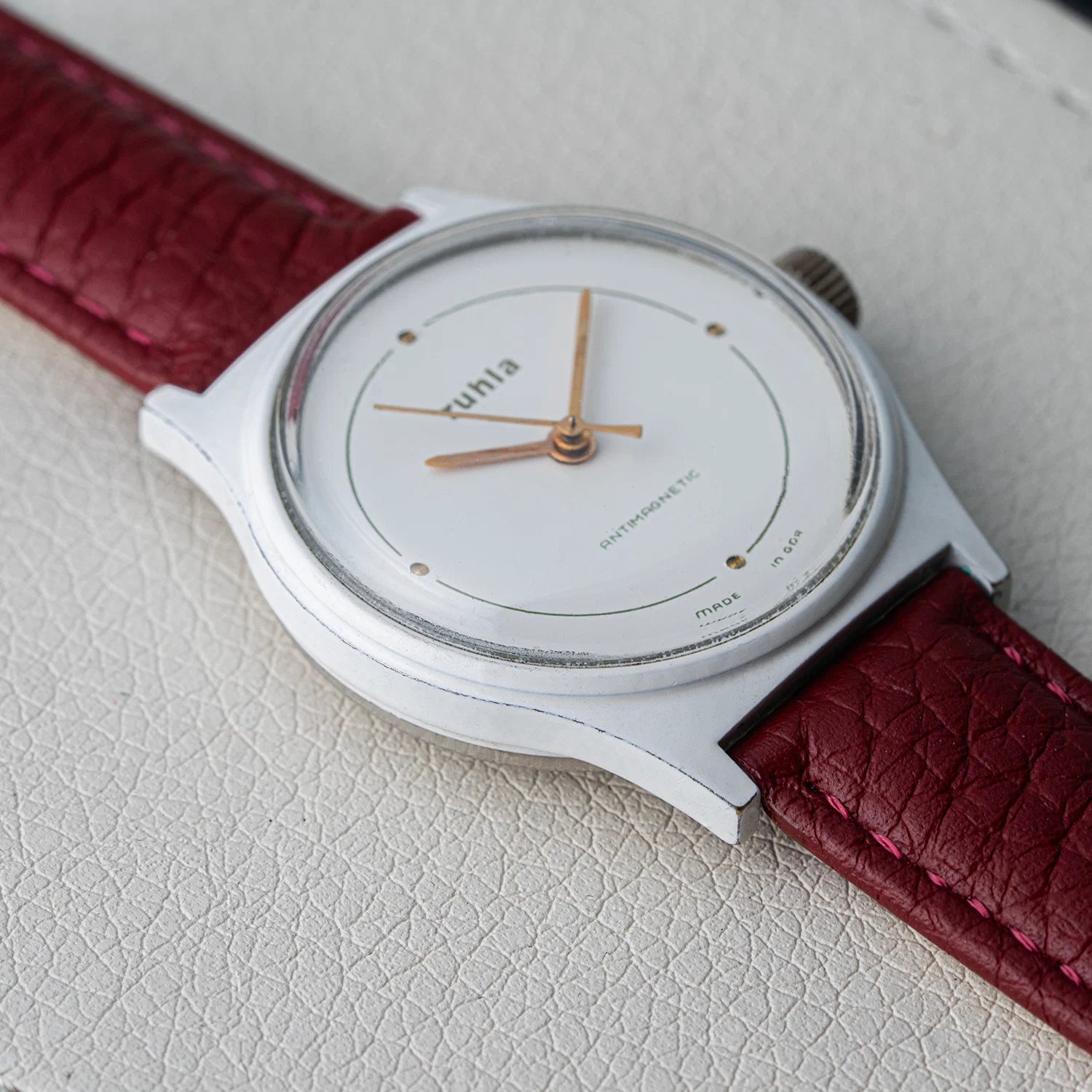 A close-up of a minimalist wristwatch Ruhla, Cal. UMF 24-42 with a silver-toned case, white dial, and burgundy textured leather strap, resting on a light textured surface.