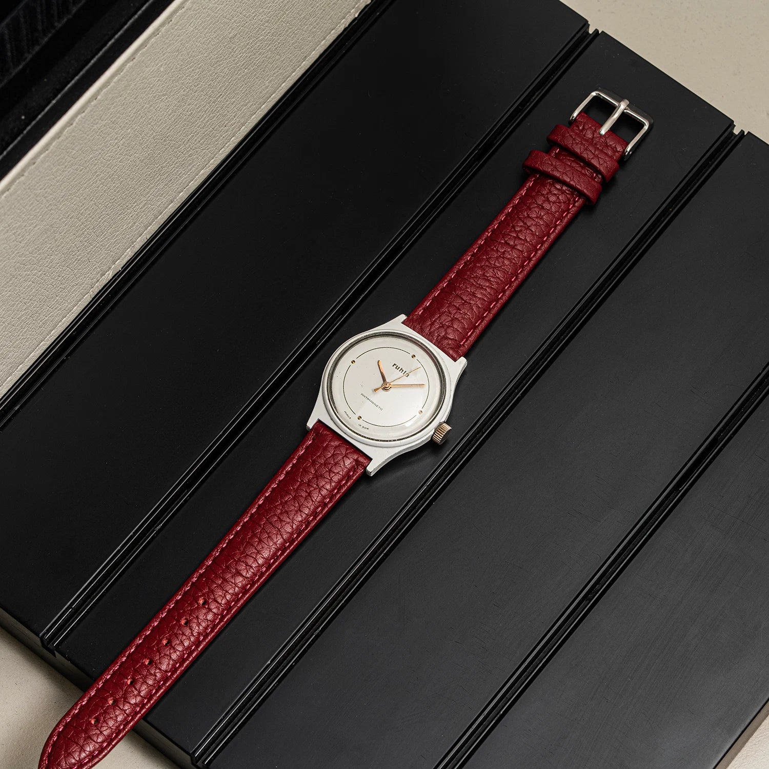 A wristwatch with a silver-toned oval case and a textured red leather strap, laid diagonally across a black rectangular surface.