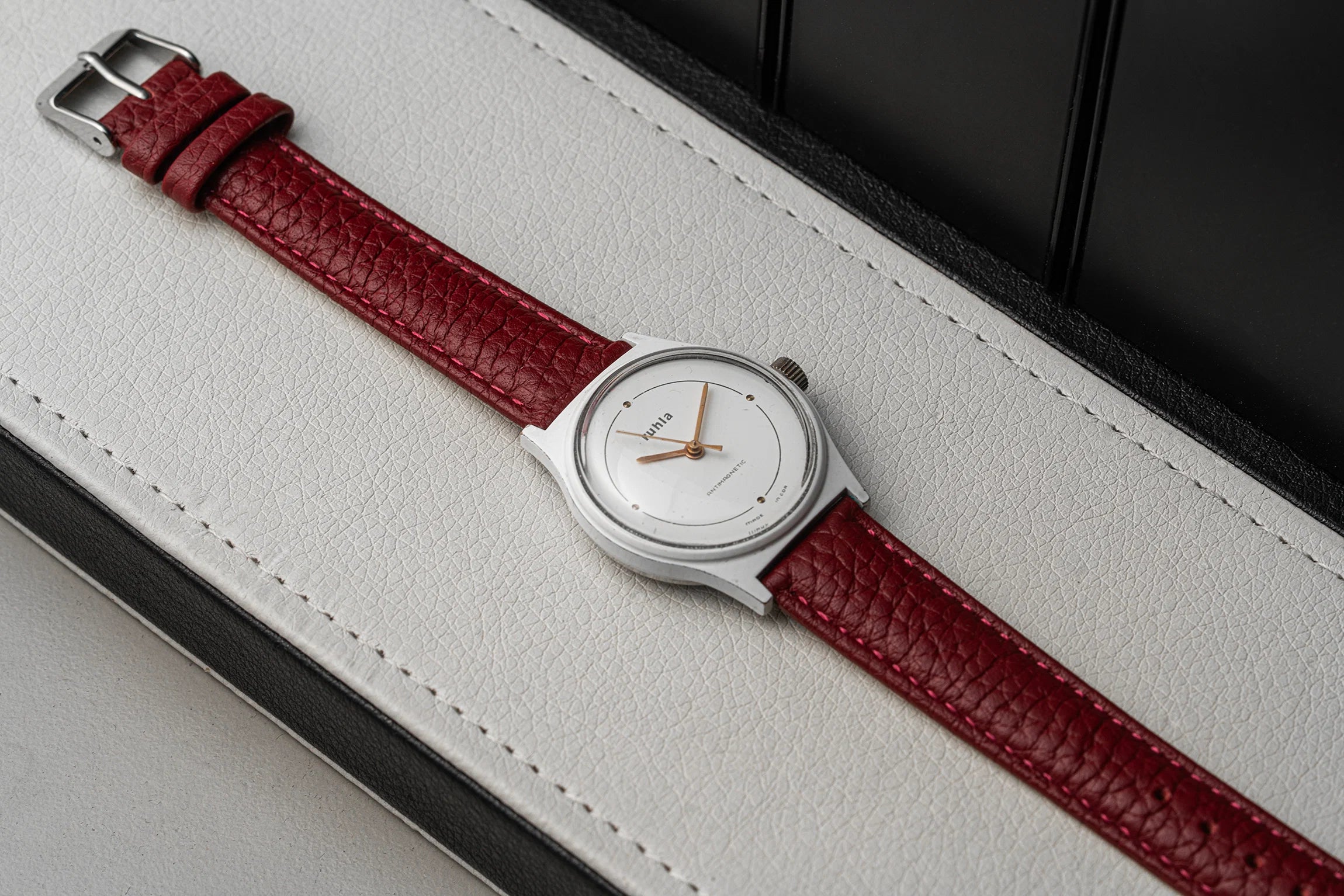A vintage-style wristwatch with a silver-toned oval case, white dial, and textured deep red leather strap, resting diagonally on a white textured leather surface with a black background.