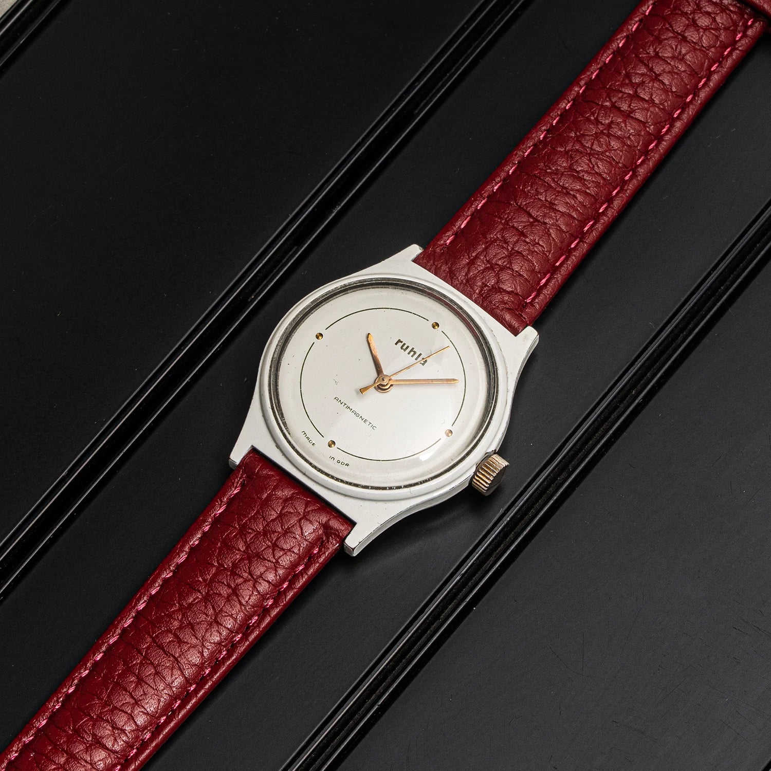 A vintage-style wristwatch Ruhla, Cal. UMF 24-42 with a silver-toned case, white dial, and textured red leather strap, photographed diagonally on a dark black background.