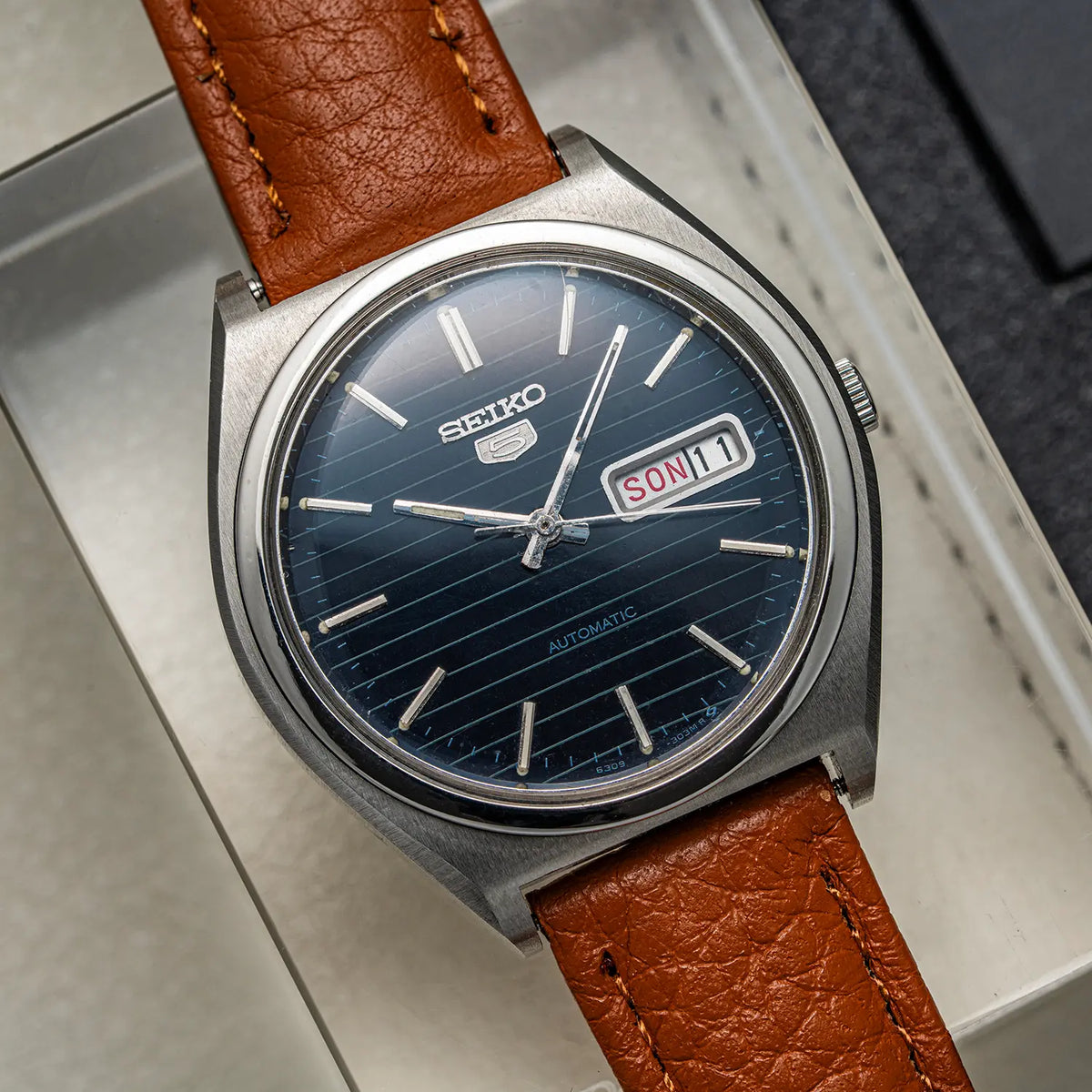 A Seiko 5, Ref. 6319-8100, Cal. 6319, Men’s Automatic, 1977 watch with a round silver-tone case, dark blue sunburst dial, and a brown leather strap, resting inside a light-colored presentation box.