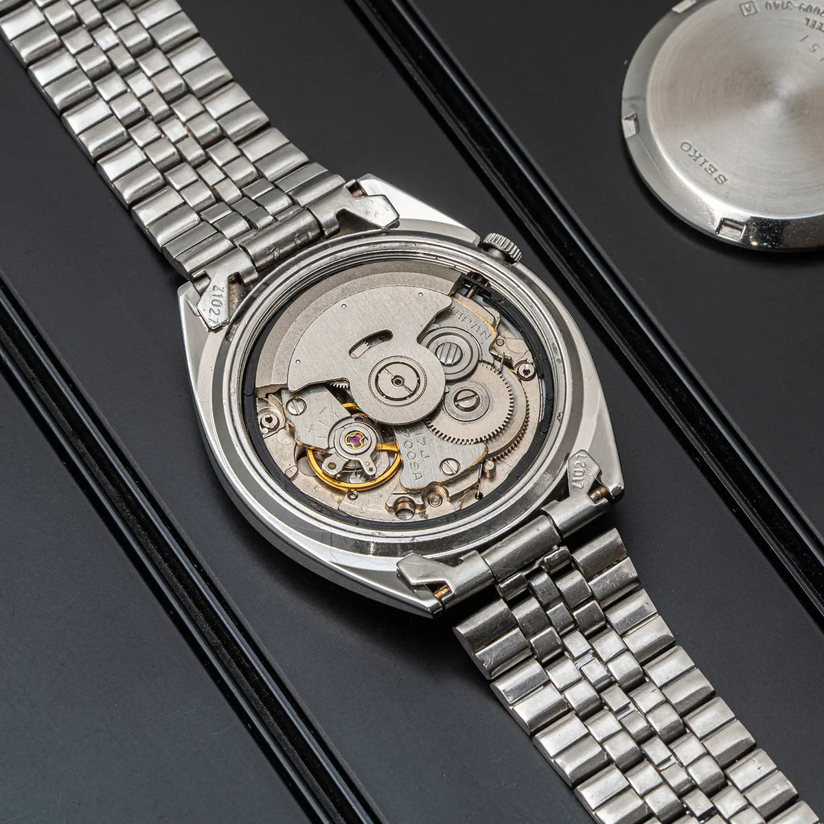 A silver stainless steel wristwatch Seiko 5, Ref. 7009-3140, Cal. 7009, Men’s Automatic, 1979 with its back open, displaying the intricate mechanical movement inside. The watch is placed on a black textured surface with a second watch case visible in the upper right corner.