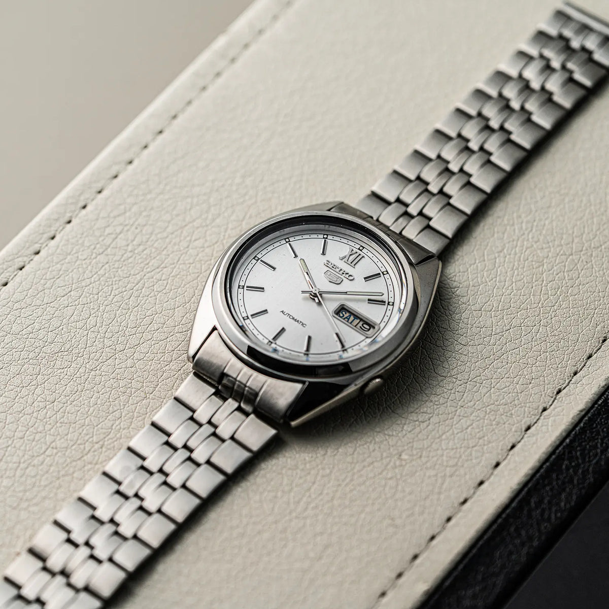 A close-up of a Seiko 5, Ref. 7009-3140, Cal. 7009, Men’s Automatic, 1979 wristwatch with a silver metal bracelet and white dial, resting diagonally on a light beige textured leather surface.