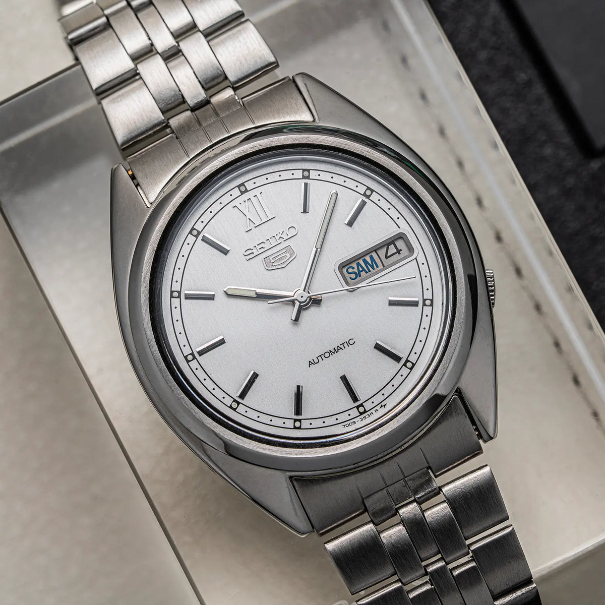 A close-up shot of a silver stainless steel wristwatch Seiko 5, Ref. 7009-3140, Cal. 7009, Men’s Automatic, 1979 with a white dial, featuring a date window and Roman numeral hour markers, resting inside a light-colored presentation box.