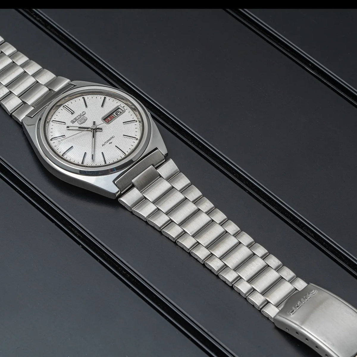 A stainless steel wristwatch Seiko 5, Ref. 7009-8750, Cal. 7009, Men’s Automatic, 1984 with a silver-toned metal bracelet and a white dial, photographed diagonally on a dark textured background.