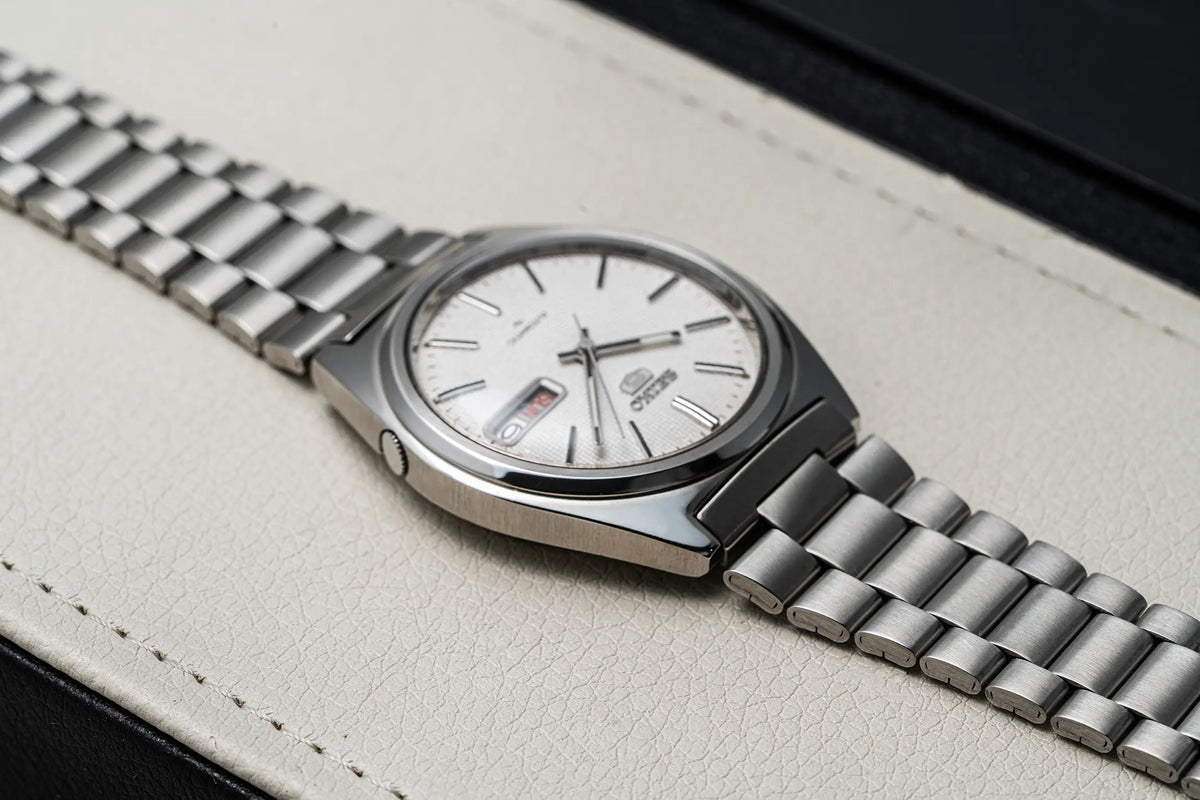 A close-up shot of a wristwatch Seiko 5, Ref. 7009-8750, Cal. 7009, Men’s Automatic, 1984 with a silver-toned metal link bracelet and a white dial, resting diagonally on a textured beige leather surface.