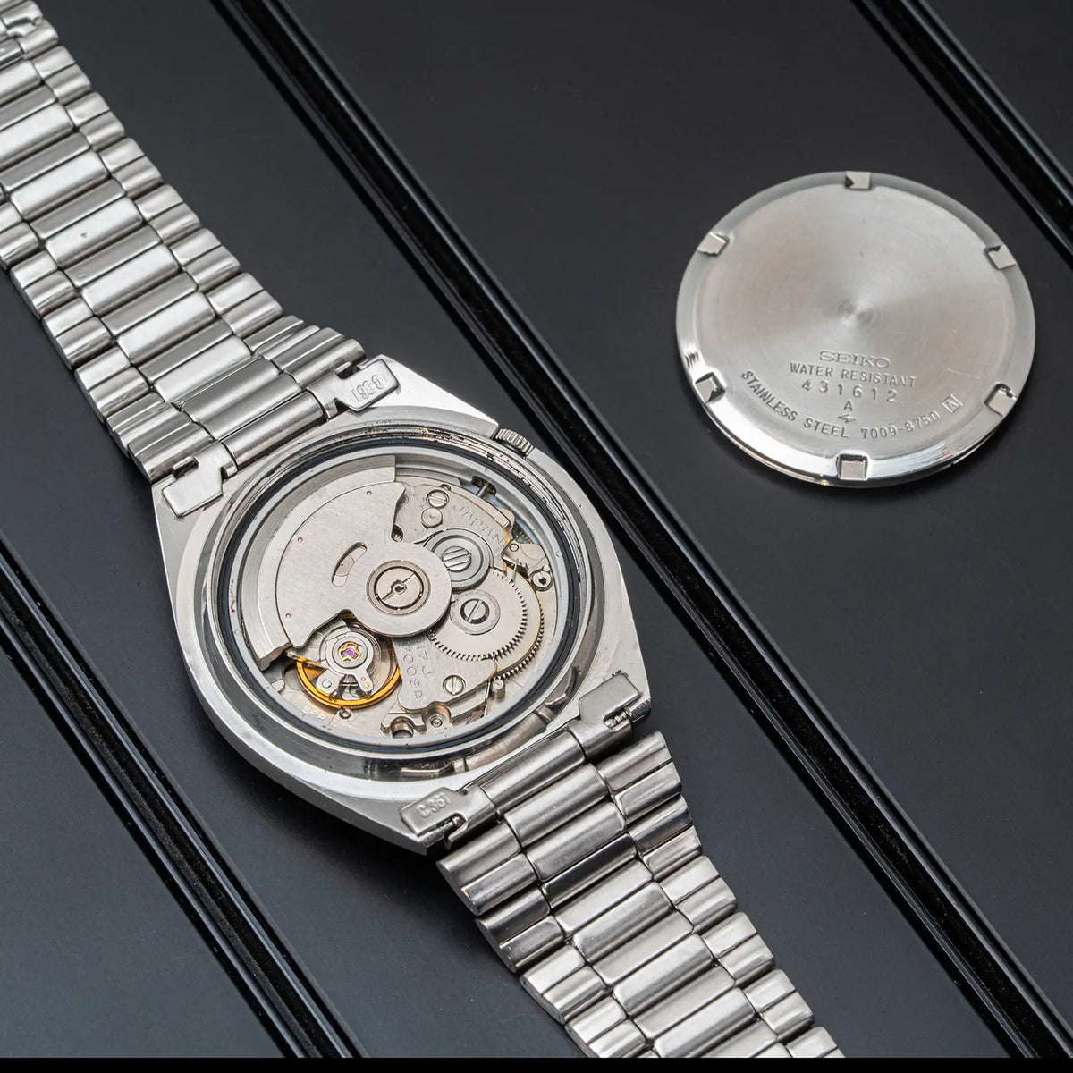 A silver stainless steel wristwatch Seiko 5, Ref. 7009-8750, Cal. 7009, Men’s Automatic, 1984 with a transparent back cover showing the intricate mechanical movement inside, displayed diagonally next to its detached circular silver case back on a dark textured background.