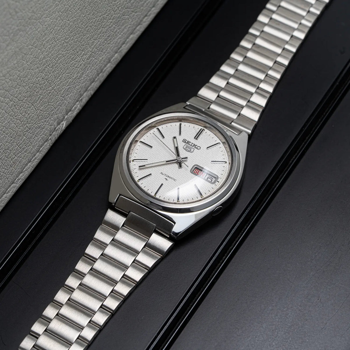 A Seiko 5, Ref. 7009-8750, Cal. 7009, Men’s Automatic, 1984 wristwatch with a silver stainless steel link bracelet and a white dial, resting diagonally on a black surface next to a gray textured leather surface.