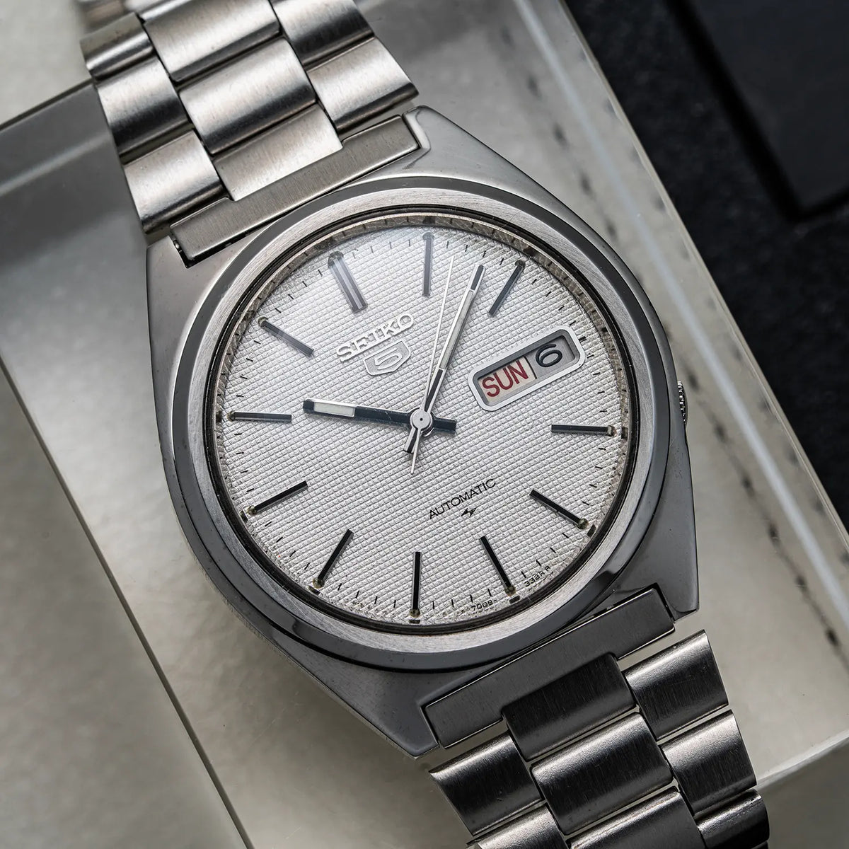 A close-up shot of a Seiko 5, Ref. 7009-8750, Cal. 7009, Men’s Automatic, 1984 automatic wristwatch with a silver-toned metal case and link bracelet, featuring a textured silver dial with black indices and a day-date window showing 'SUN'.