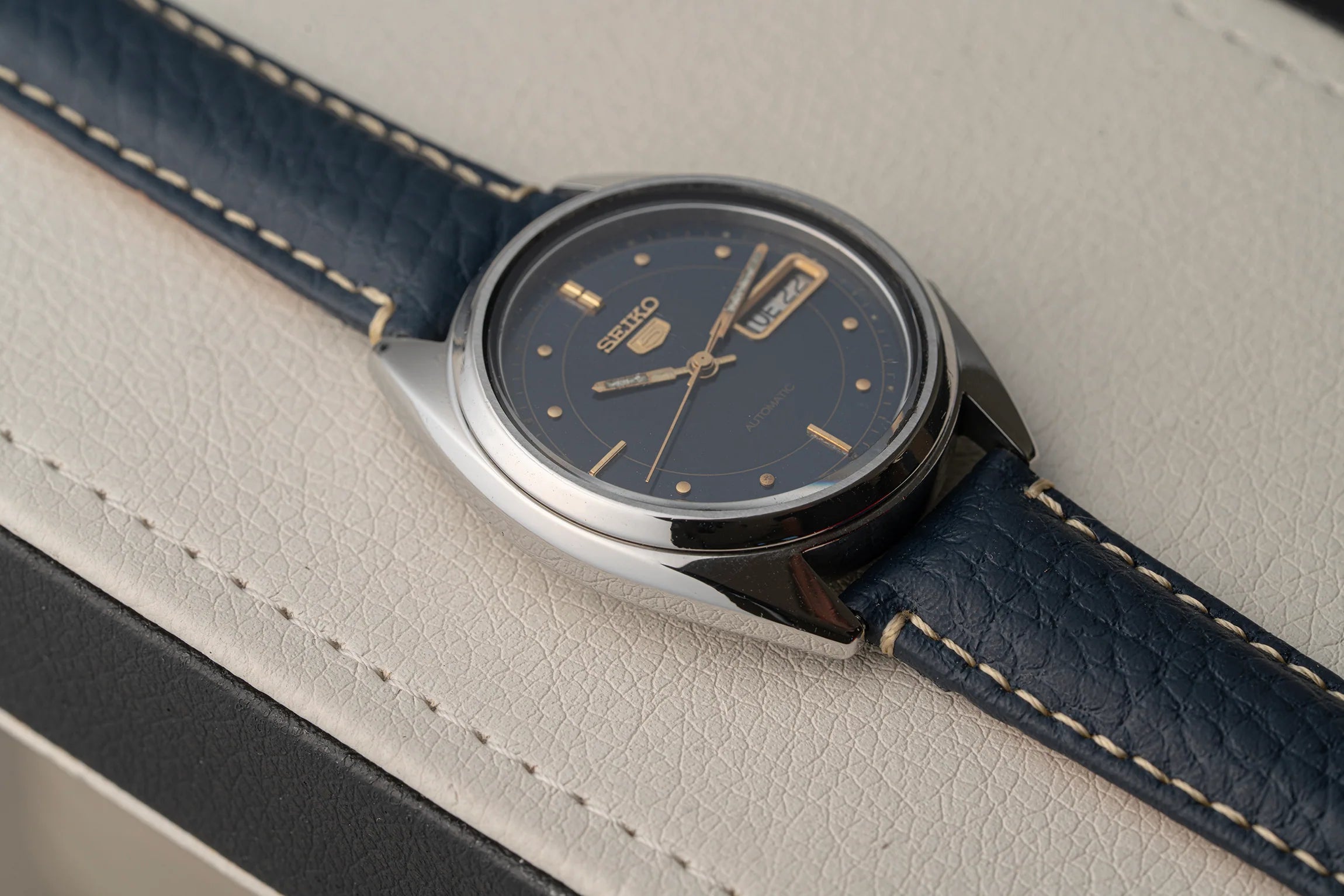 A close-up of a Seiko 5 7009-3040 wristwatch with a dark blue dial and a dark blue leather strap with light contrast stitching, resting on a textured light beige leather surface.