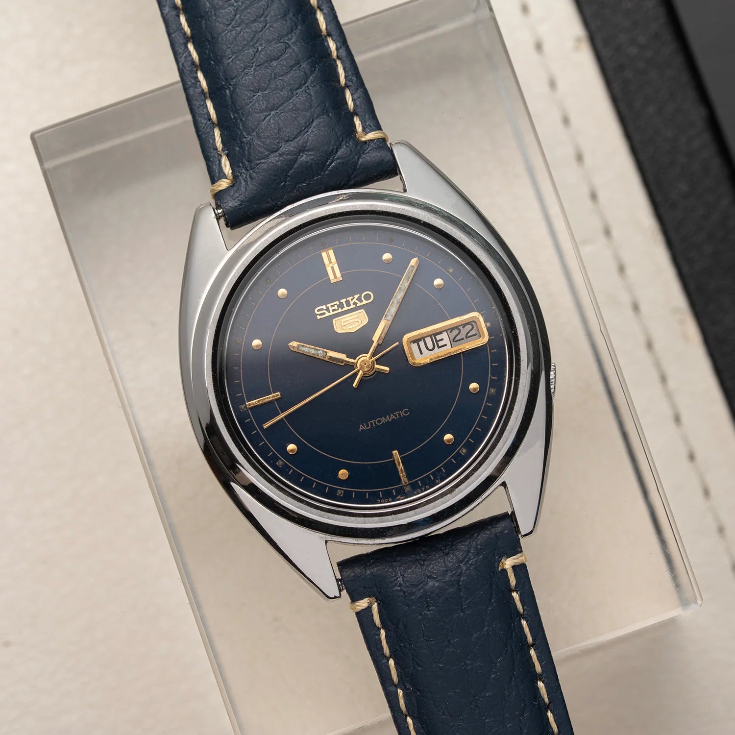 A vintage Seiko 5 7009-3040 automatic watch with a dark blue leather strap and a matching dark blue dial, featuring gold-tone hands, markers, and a date window. The watch is displayed inside a clear plastic presentation box on a light beige surface.