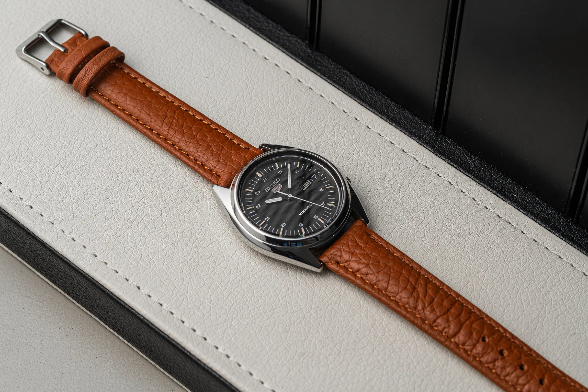A wristwatch Seiko 5 7009-3040 with a brown textured leather strap and a dark round dial, resting diagonally on a light beige leather surface with a black background.
