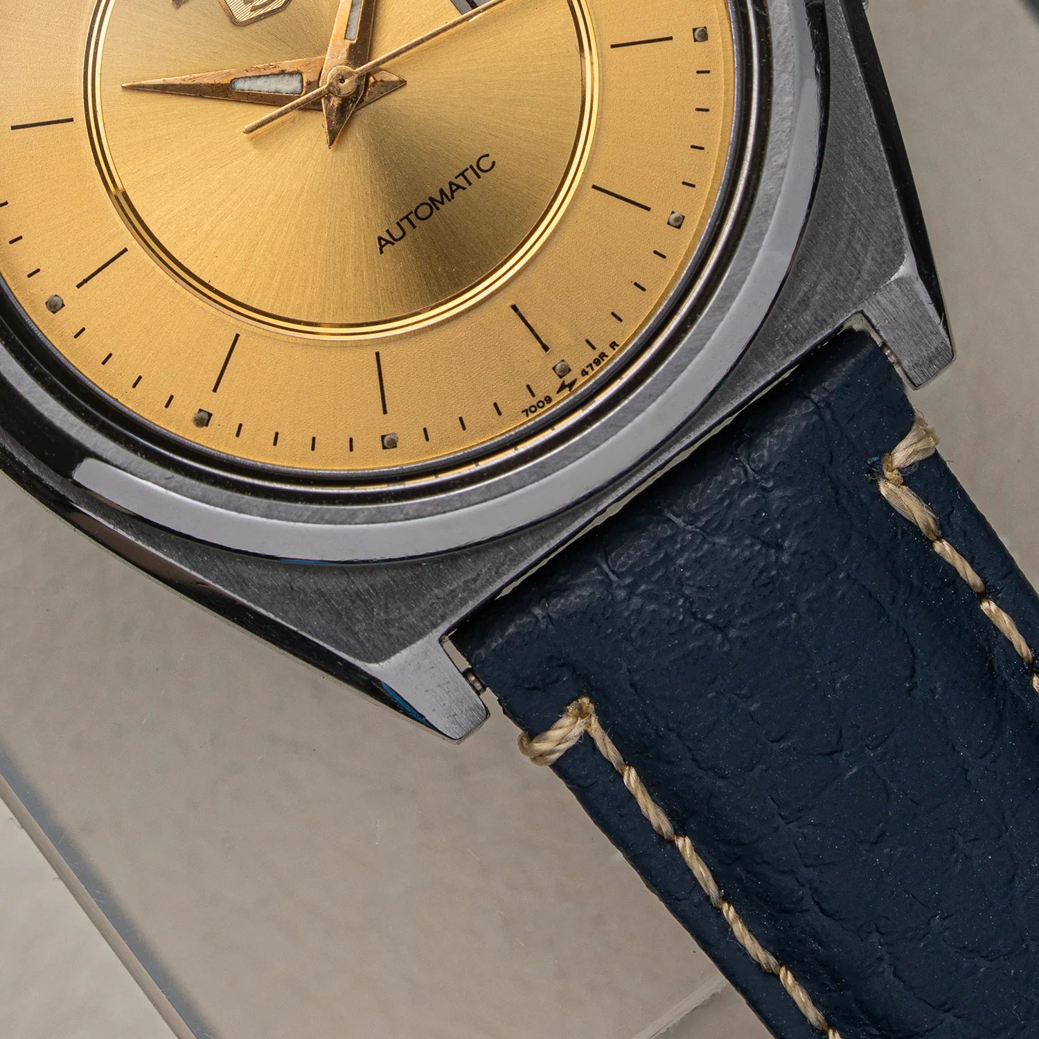 Close-up of a wristwatch Seiko 5 7009-876A with a brushed silver-tone case, a gold dial, and a dark blue leather strap with light stitching.