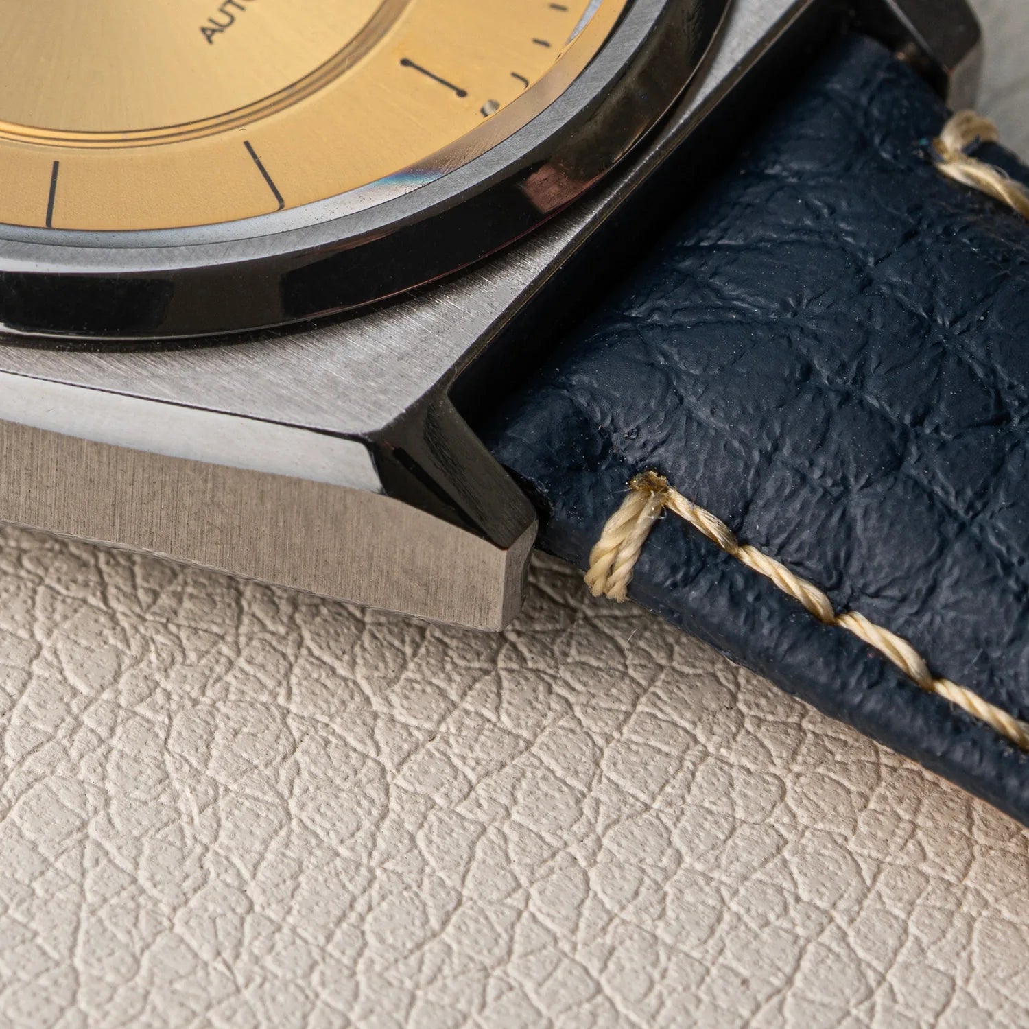 Close-up of a wristwatch Seiko 5 7009-876A showing a brushed silver metal case, a gold-toned dial, and a dark navy textured leather strap with cream stitching, resting on a light beige textured surface.