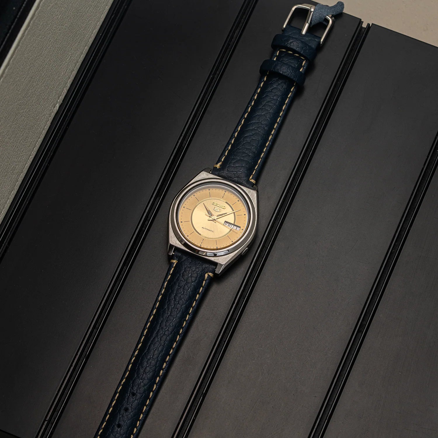 A vintage-style wristwatch Seiko 5 7009-876A with a round silver-toned case, a gold dial, and a dark blue textured leather strap with light contrast stitching, displayed diagonally on a dark textured background.