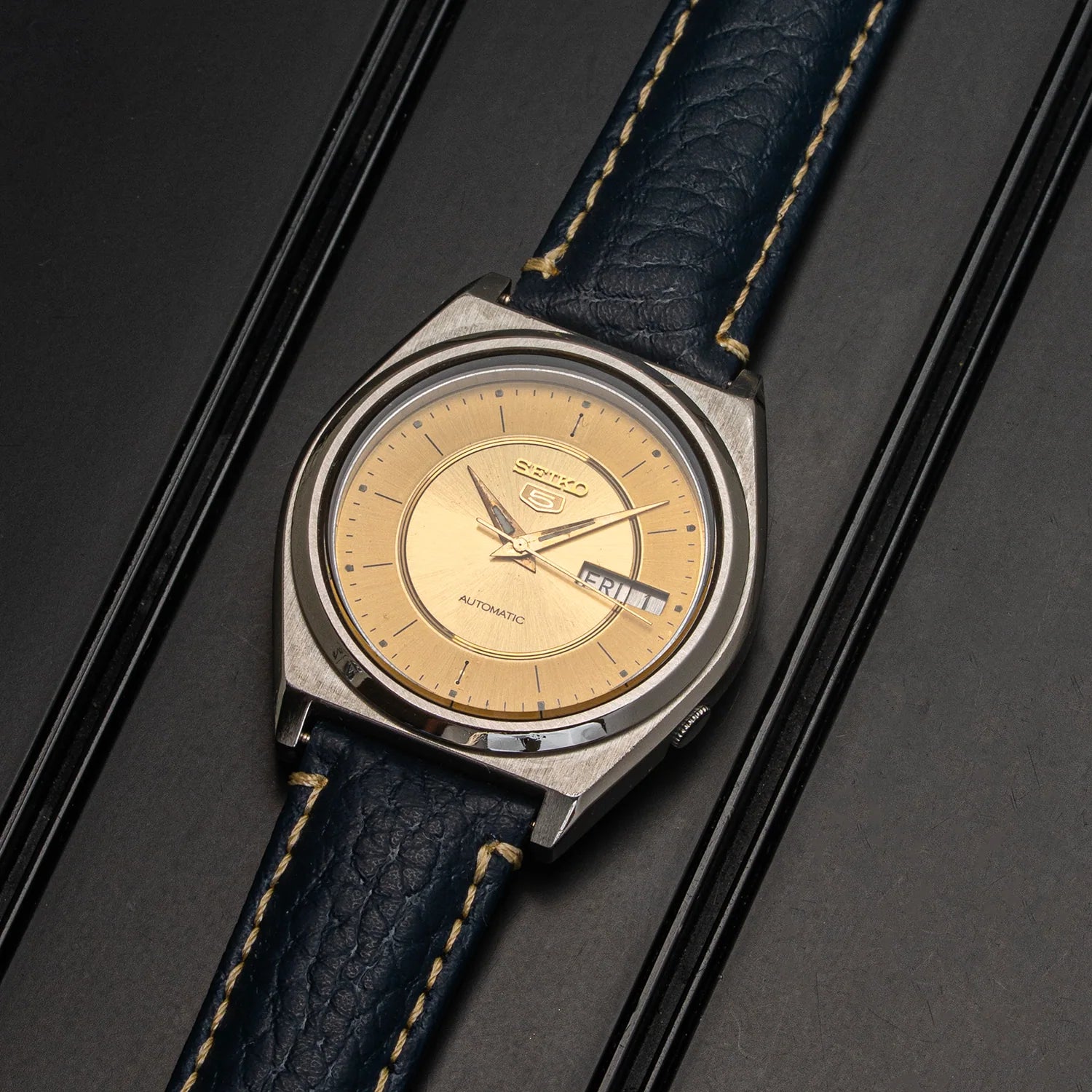 A vintage Seiko watch Seiko 5 7009-876A with a round silver-tone metal case, a beige dial, and a dark blue leather strap with light tan stitching, shown on a dark textured background.