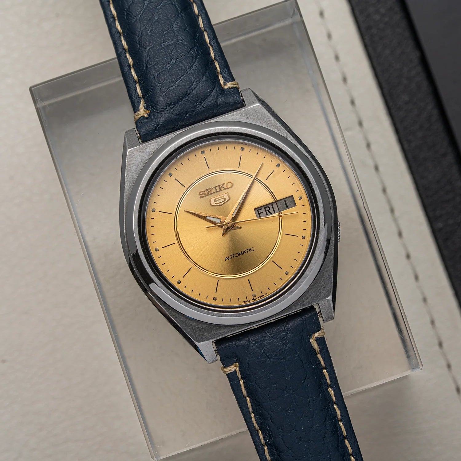 A vintage Seiko 5 7009-876A watch with a gold dial and a dark blue leather strap, displayed on a clear acrylic stand.
