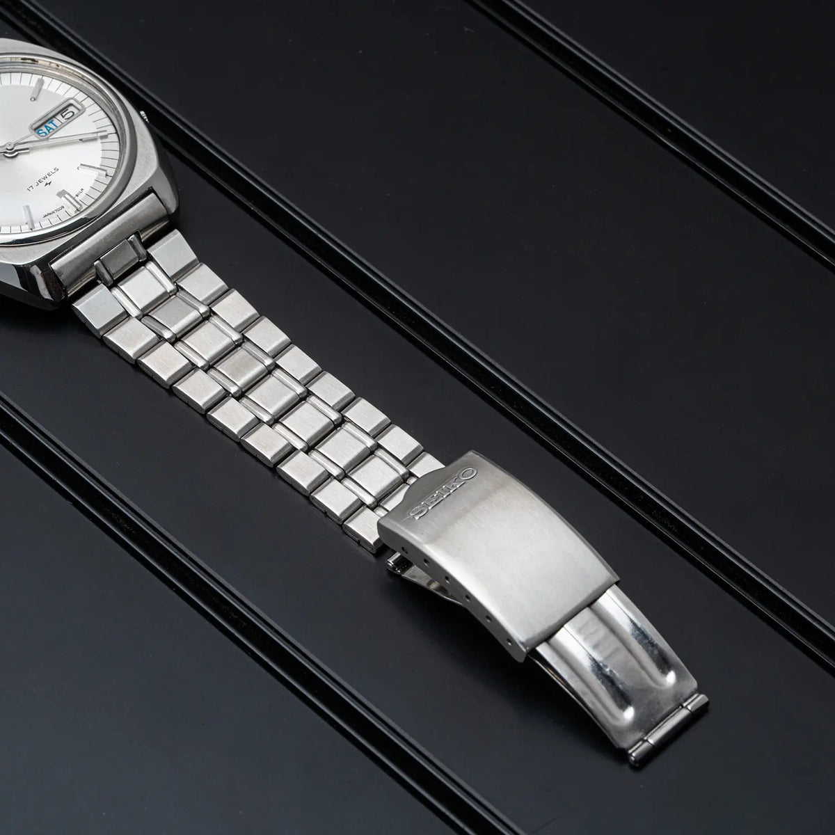 A close-up of a Seiko, Ref. 7009-8100, Cal. 7009, Men’s Automatic, 1977 stainless steel watch band with a silver-toned clasp and white dial, laid diagonally on a dark textured surface.